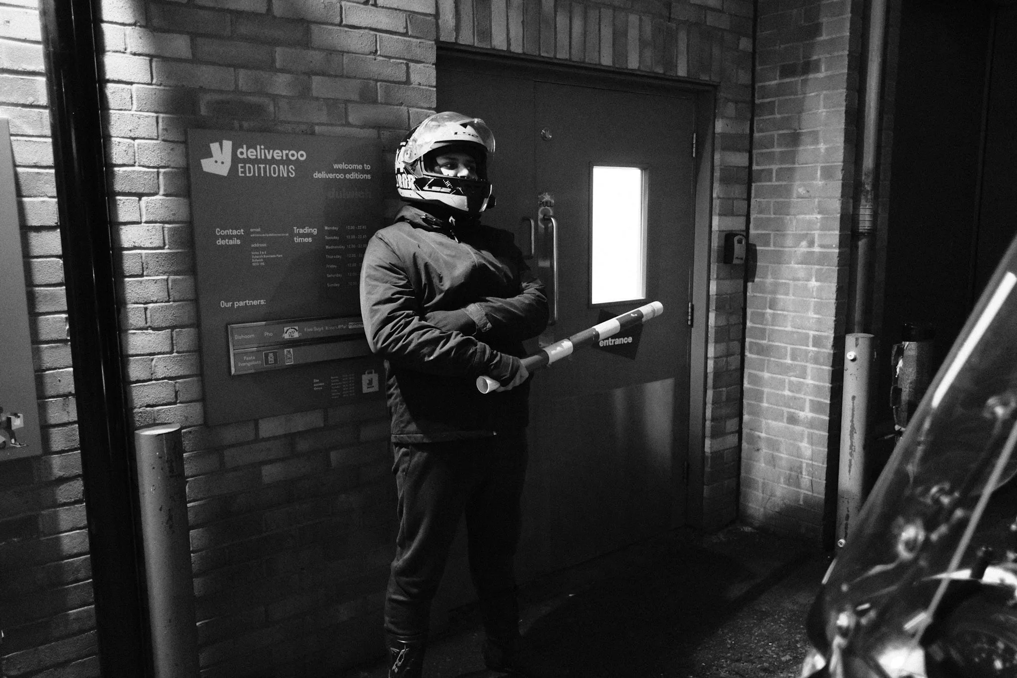 A courier in a full-face helmet stands with arms folded at the entrance of a Deliveroo Editions unit at night, a raised barrier arm across the doorway beside a wall sign listing the site's trading times and restaurant partners.