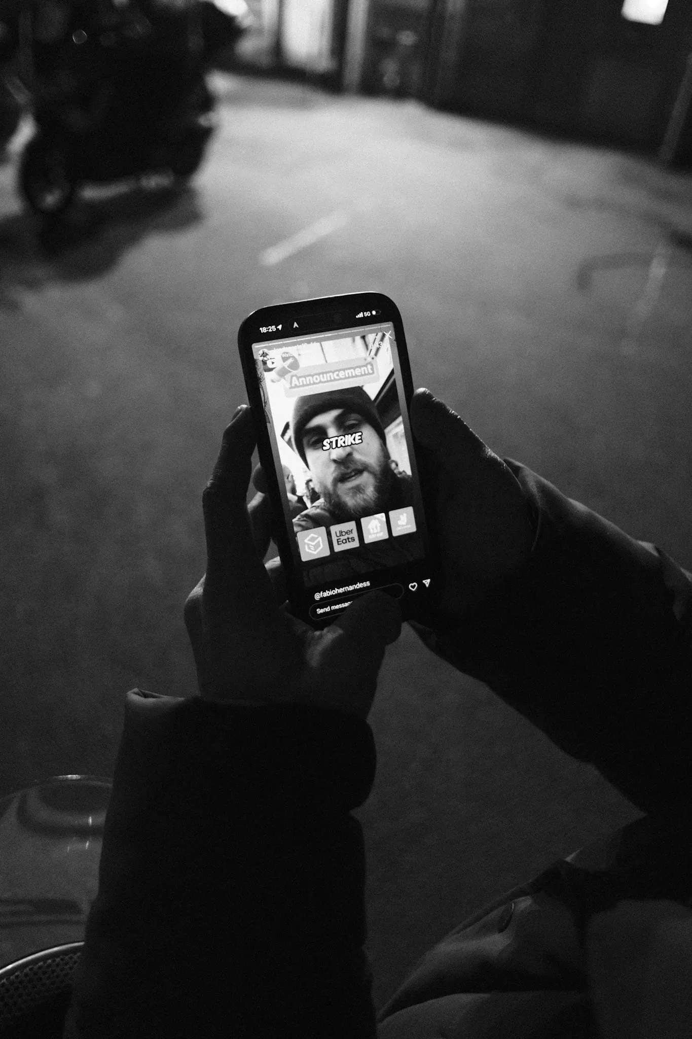 Gloved hands hold up a phone in a dark yard. On the screen an Instagram story headed Announcement shows a courier speaking to camera with the word STRIKE across his face, the icons of four delivery apps in a row beneath him.