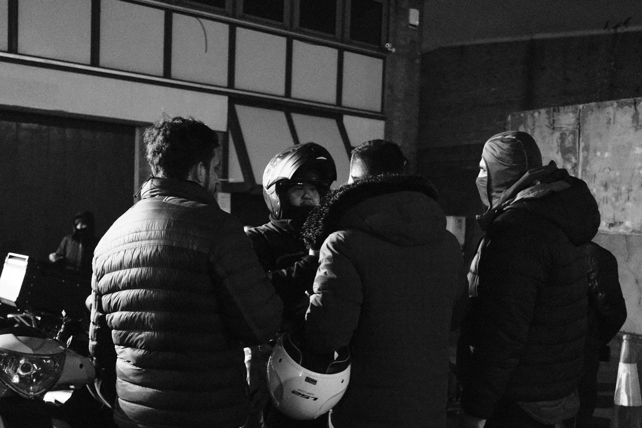 Five couriers in winter coats and helmets stand close together talking in the dark outside an industrial unit, a scooter and a traffic cone at the edges of the frame.