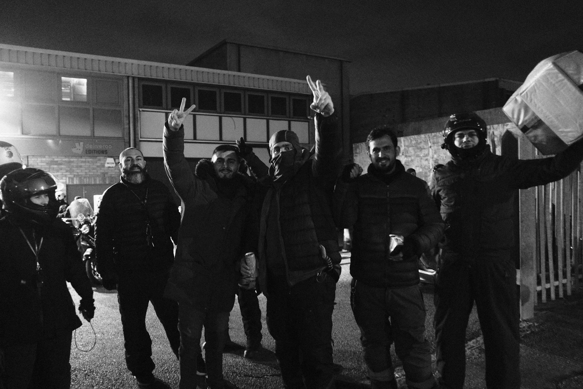 A line of couriers outside the Deliveroo Editions unit at night, several with arms raised in V-signs and one holding a delivery box above his head, all looking into the camera.