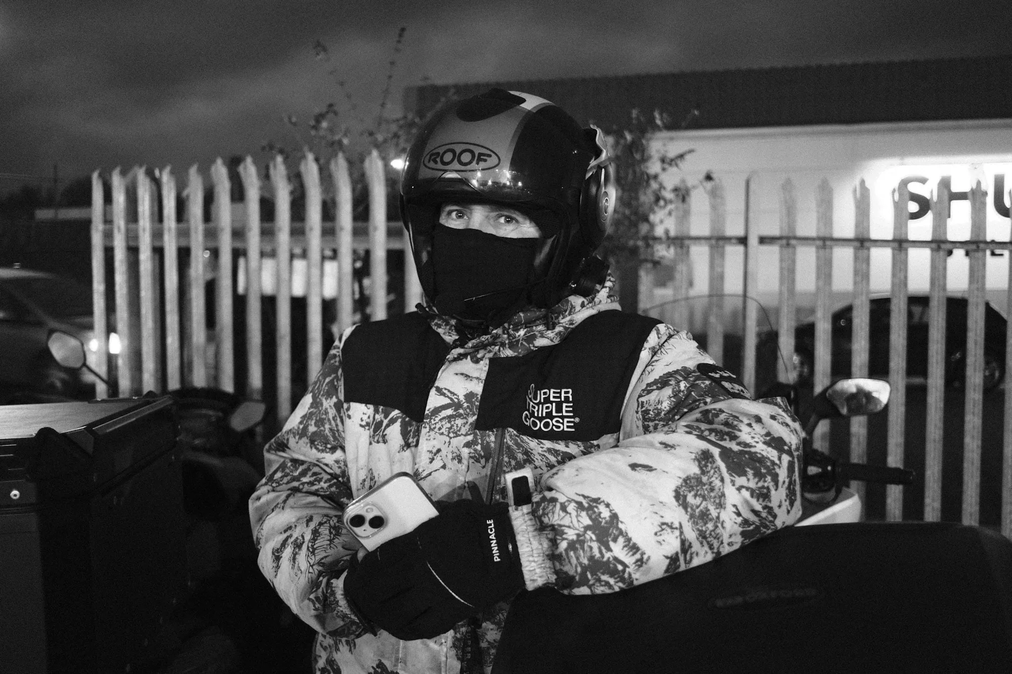 A courier in a patterned puffer jacket and open-face helmet stands with arms folded and a phone in one hand, a spiked steel fence and a lit self-storage sign behind.