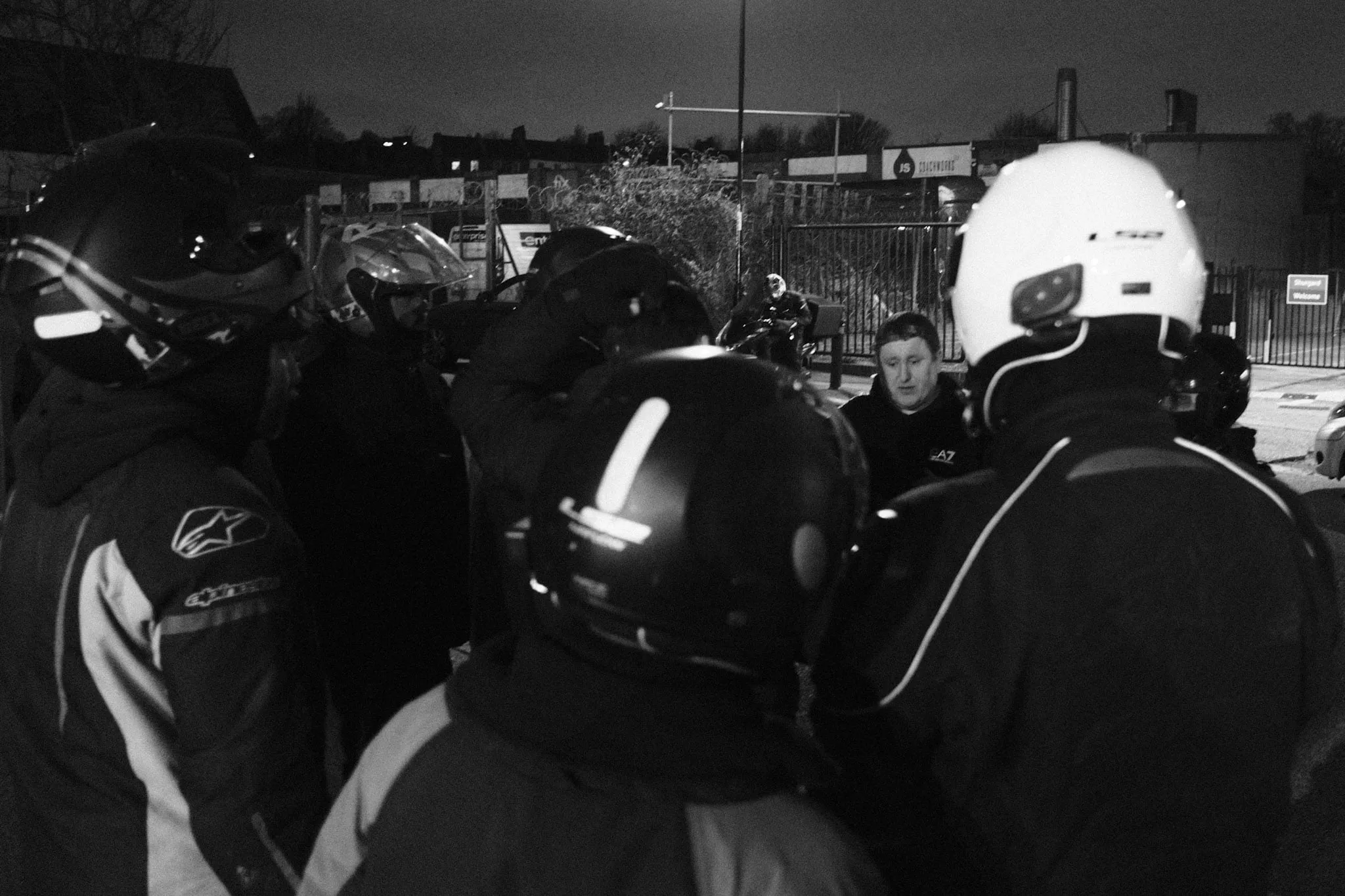 A ring of couriers seen from behind, helmets still on, listening to a man in a dark jacket standing facing them at the centre of the group.