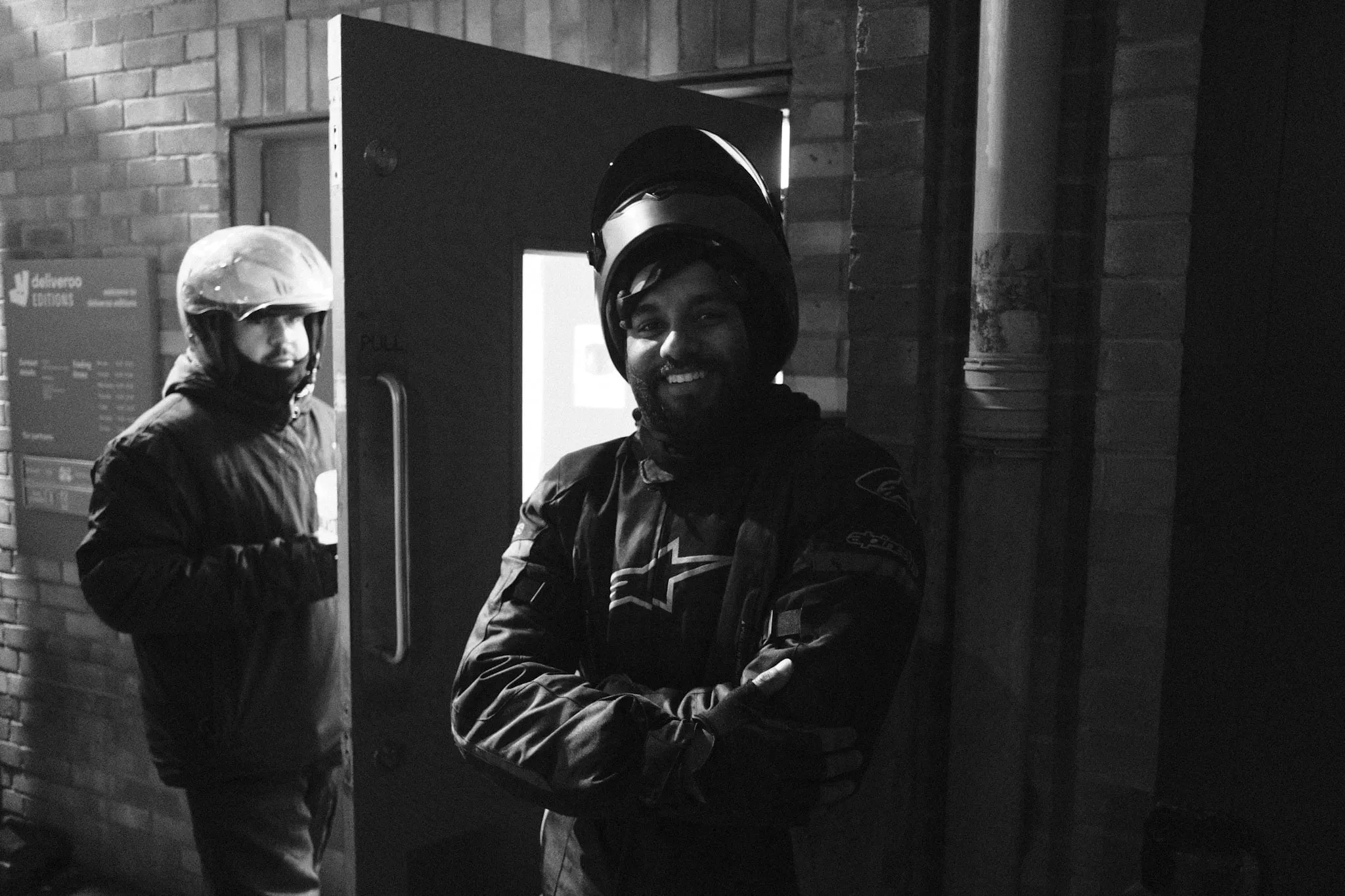 Two couriers at the Deliveroo Editions door, the one nearest the camera in an armoured jacket and helmet, arms folded, grinning.