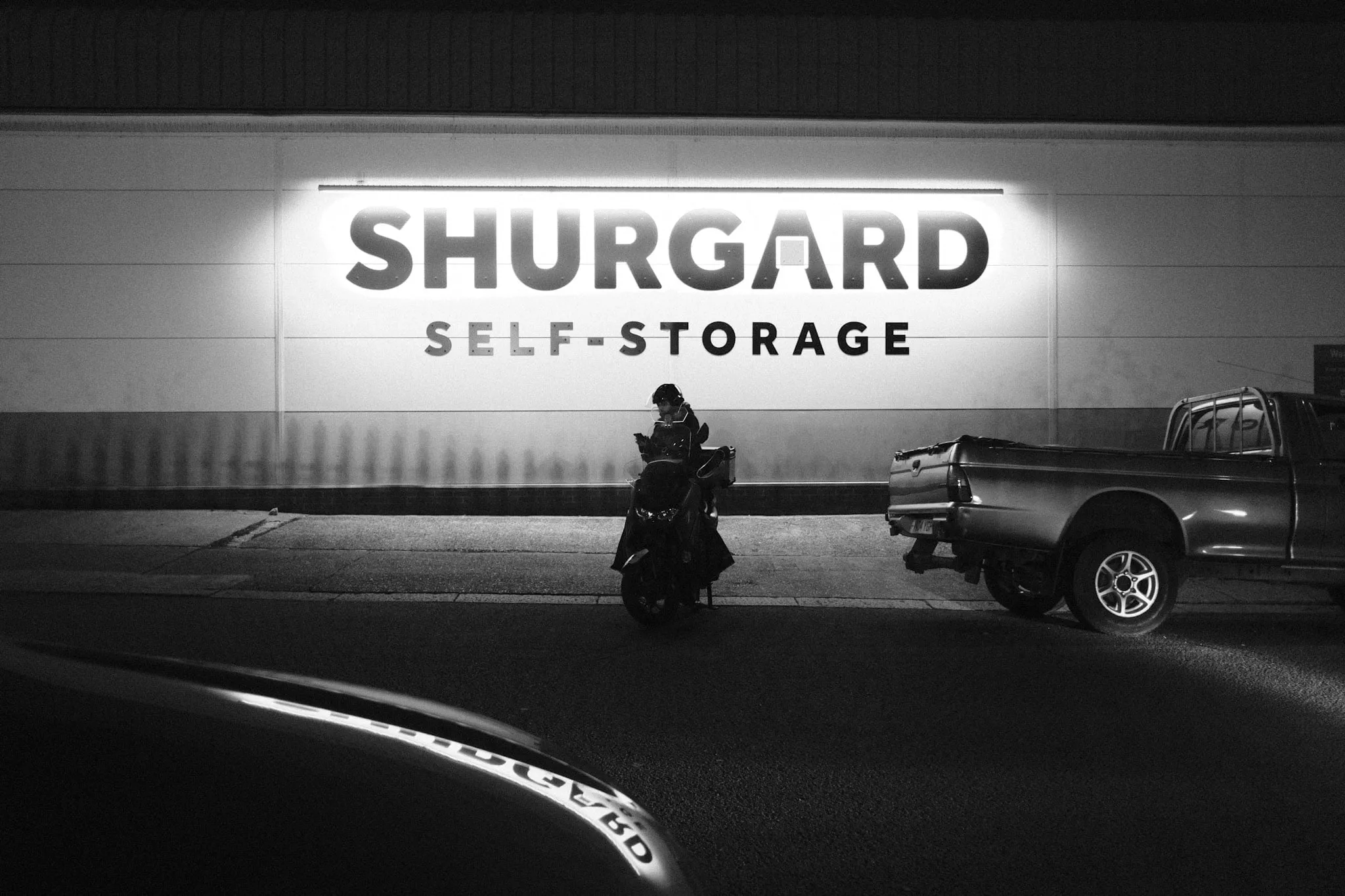 A courier sits on a scooter checking a phone, dwarfed by an illuminated Shurgard Self-Storage sign filling the wall above.