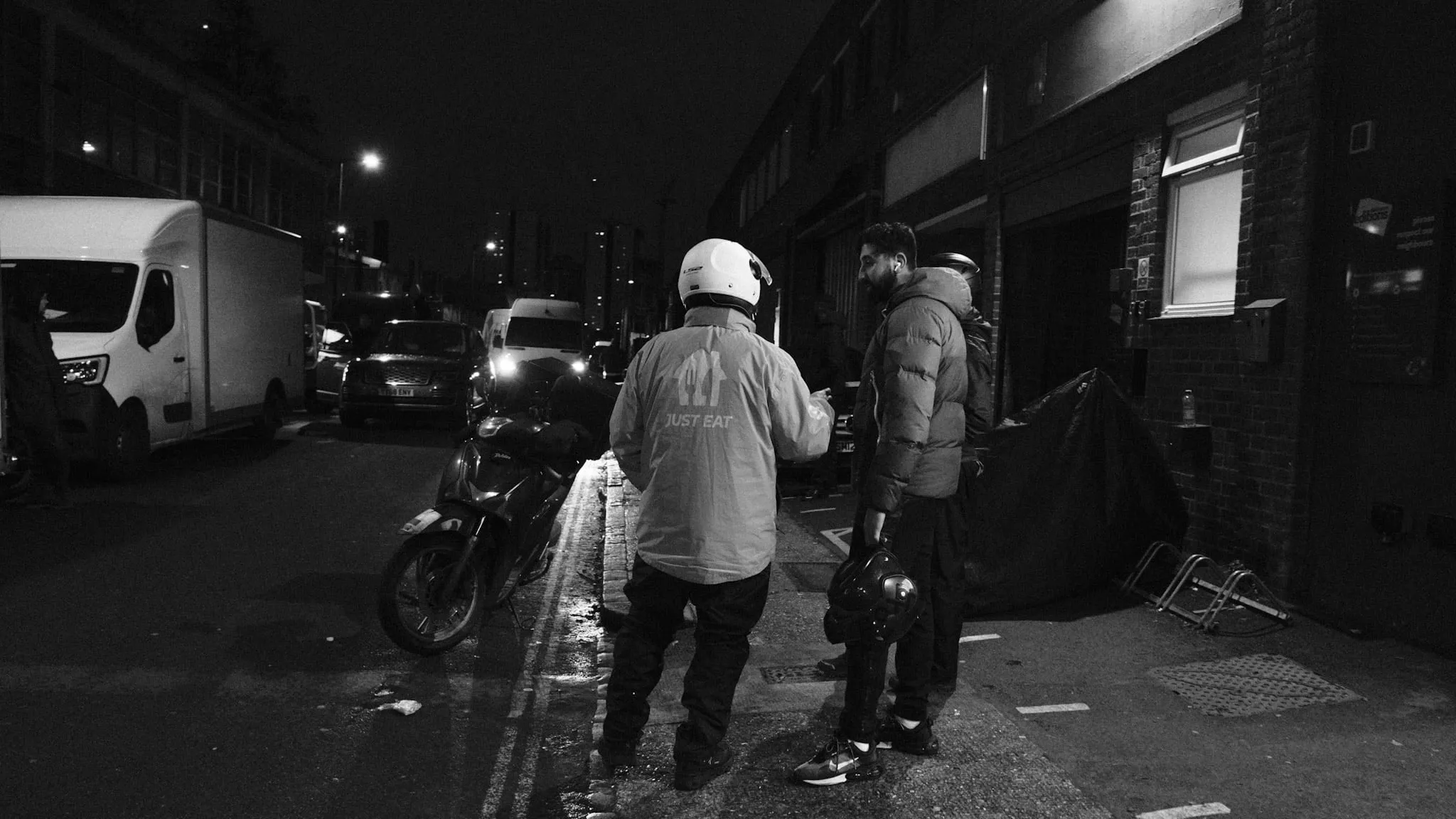 A courier in a Just Eat jacket walks away up a dark street towards two others, a scooter parked at the kerb.