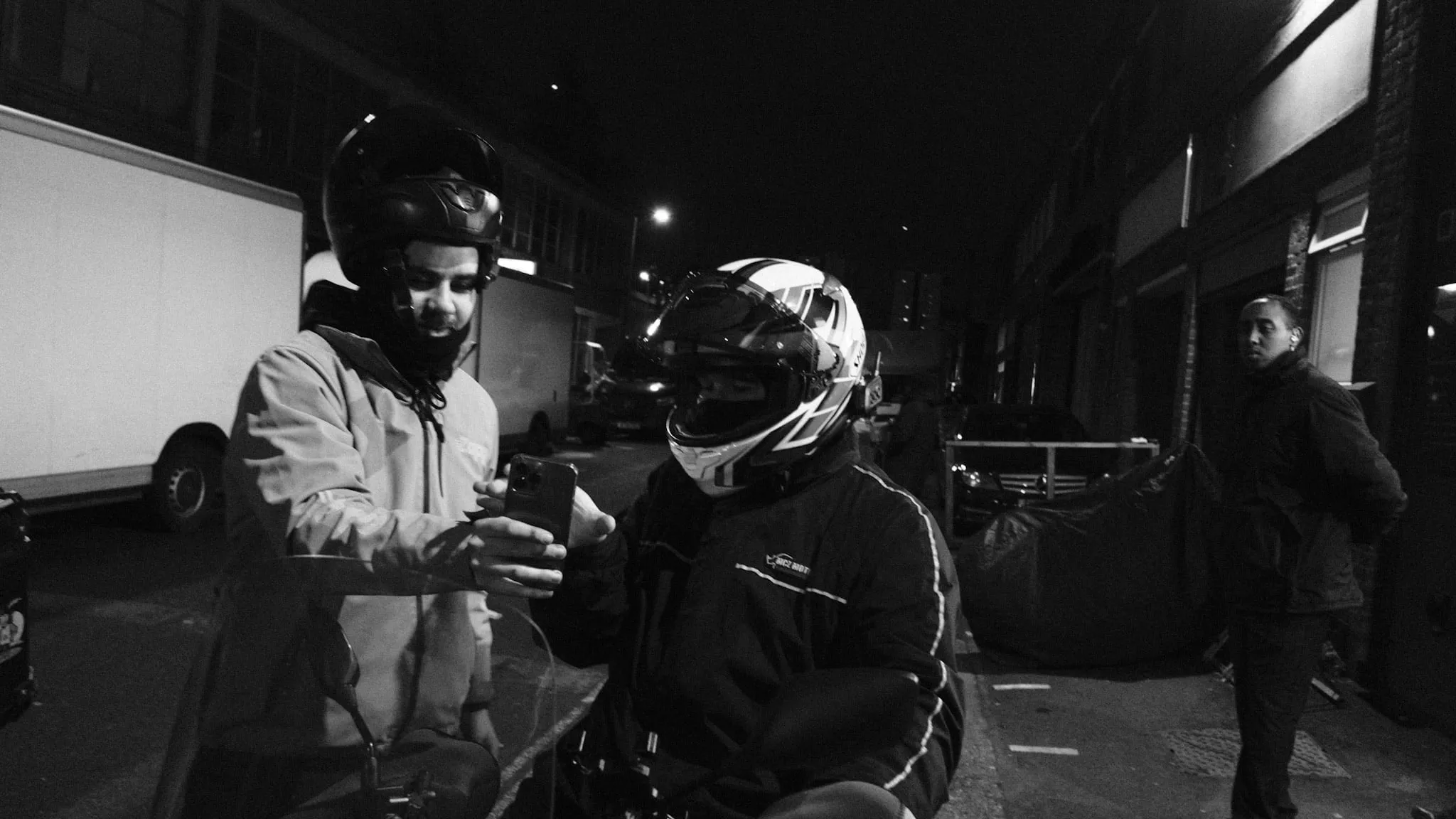 Two couriers on scooters at night, one holding out his phone for the other, in a full-face helmet, to read.