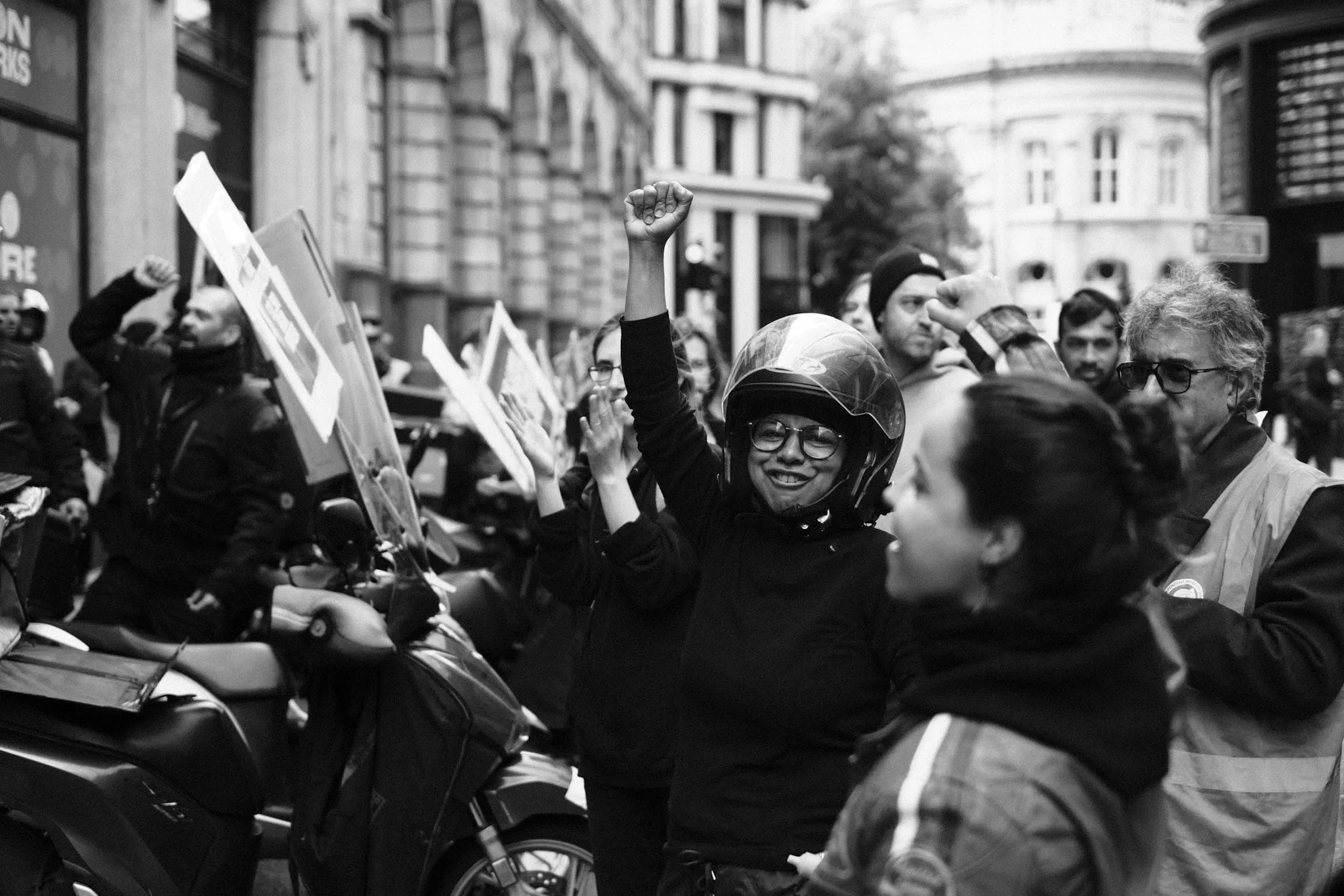 A courier in a helmet and glasses raises a clenched fist and laughs in a City street, surrounded by others with fists and placards raised, scooters lined along the kerb.