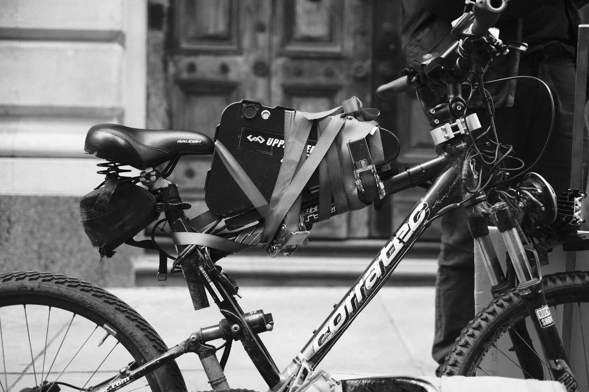 A battered mountain bike at the kerb with a courier's insulated delivery box strapped to the rack behind the saddle with webbing.