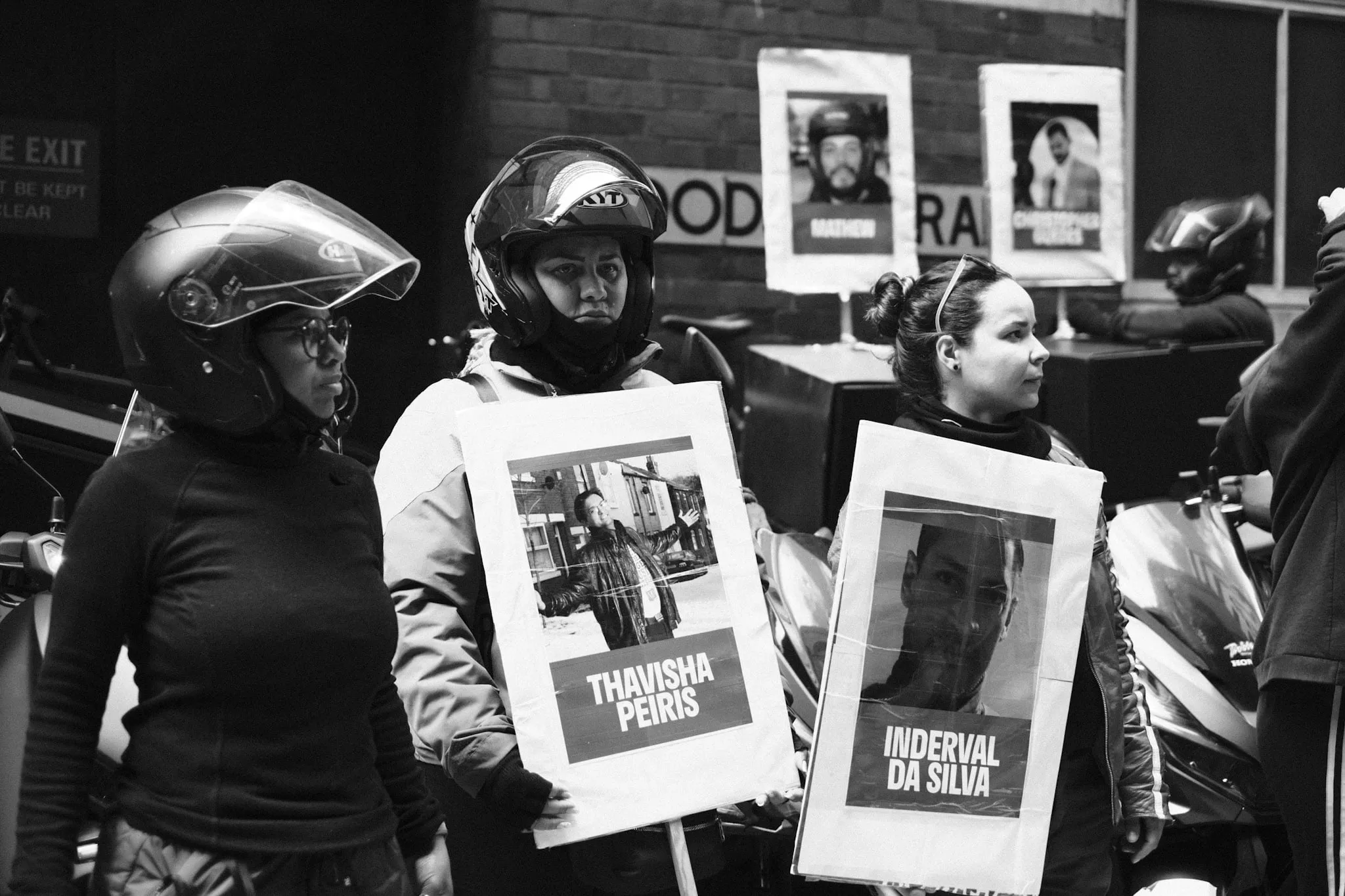Couriers stand holding placards that carry photographs and names of riders killed at work, more of the same portraits mounted on stands behind them.