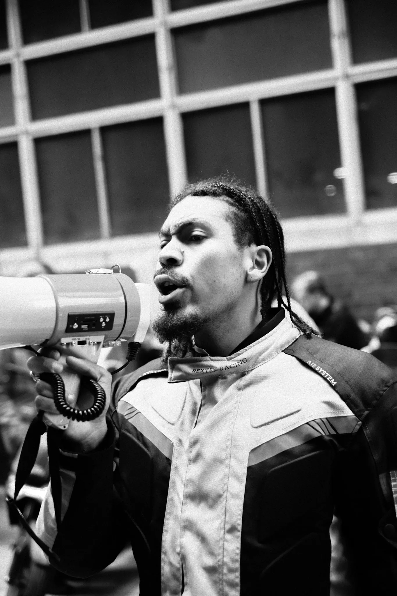 A young courier in a motorcycle jacket speaks into a megaphone, jaw set, the crowd out of focus behind him.