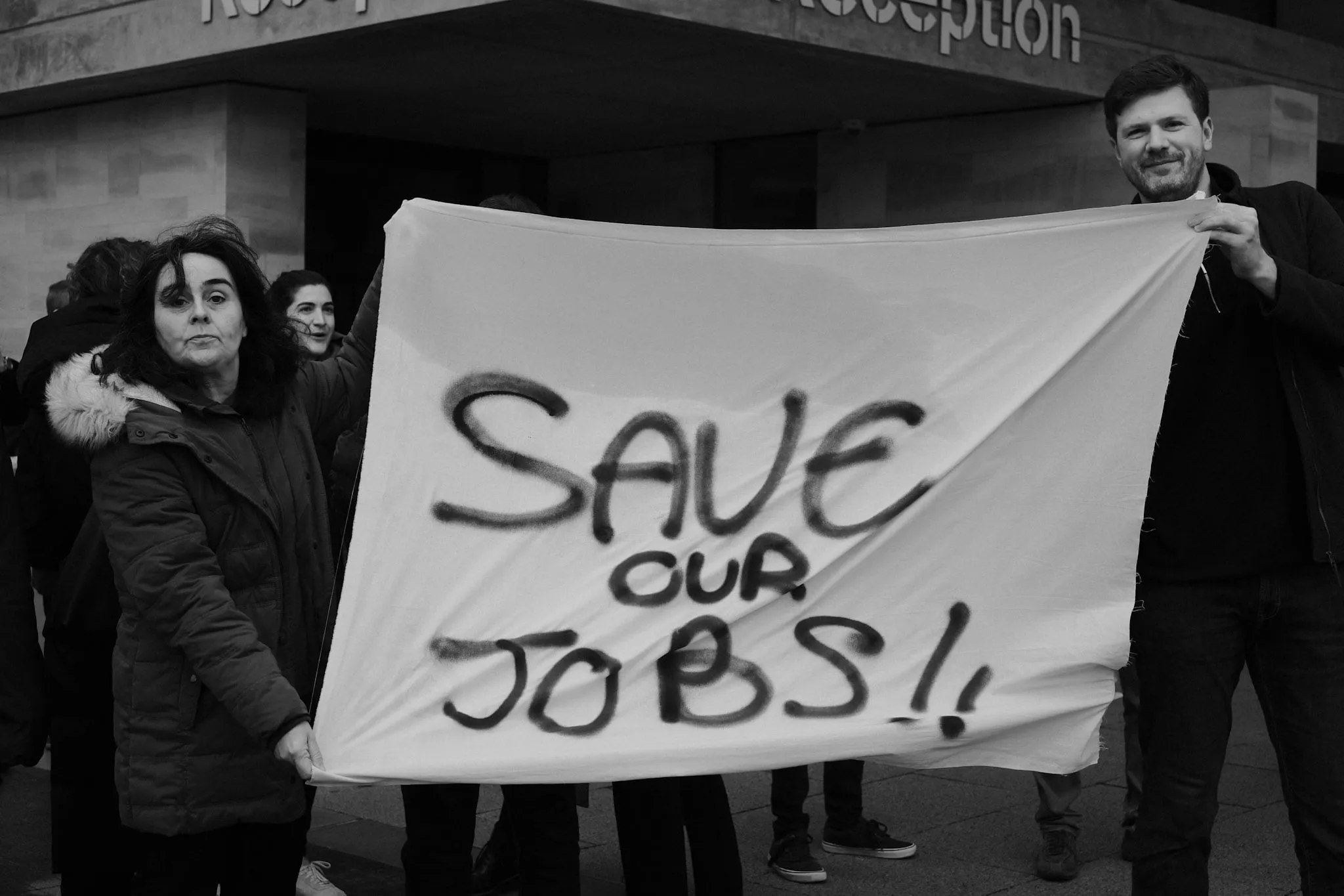 Two people hold a bedsheet banner with SAVE OUR JOBS!! spray-painted across it, standing outside a university reception entrance.