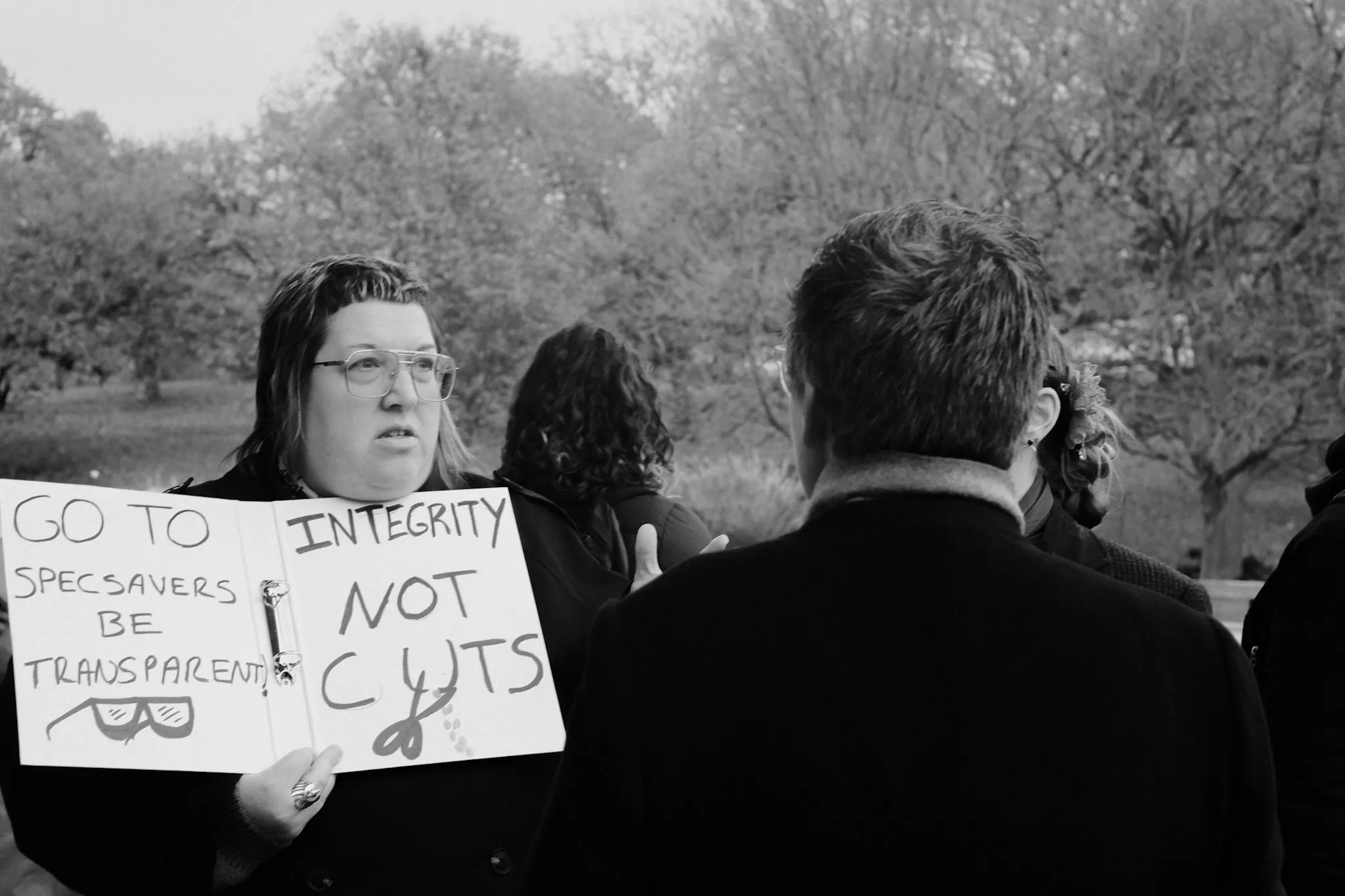 A picket holds up a ring binder with handwritten signs taped inside — GO TO SPECSAVERS, BE TRANSPARENT and INTEGRITY NOT CUTS — while others talk around her.