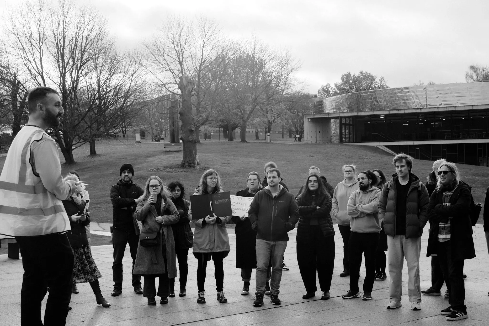 A speaker in a hi-vis vest addresses a crowd of about twenty gathered on a paved campus square, a low teaching block and bare winter trees behind them.