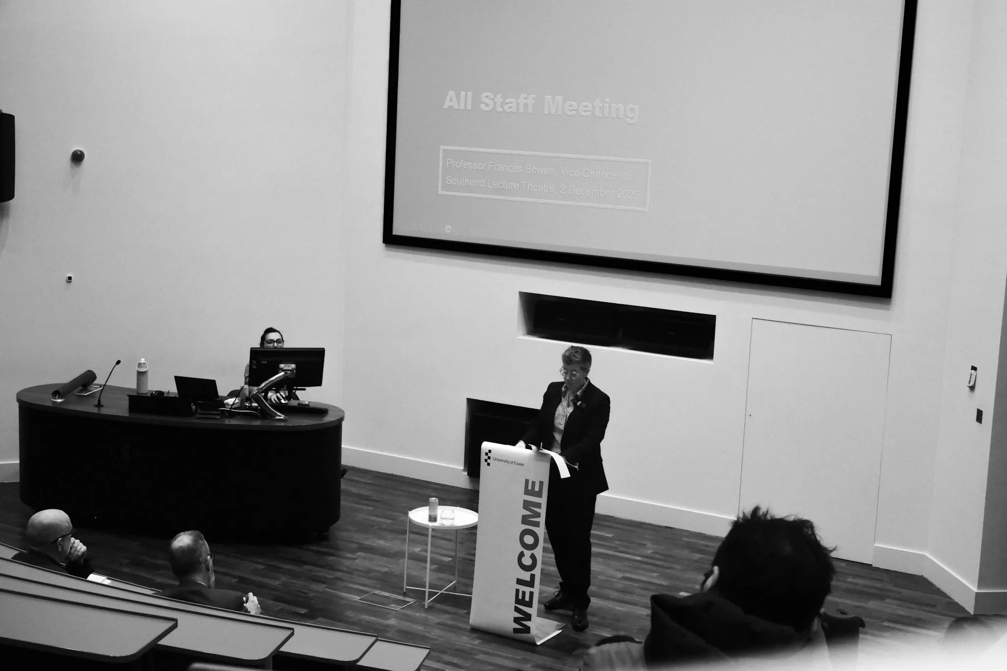 A lecture theatre seen from the back rows: the Vice-Chancellor stands at a lectern beside a screen headed All Staff Meeting, a handful of people in the seats below.