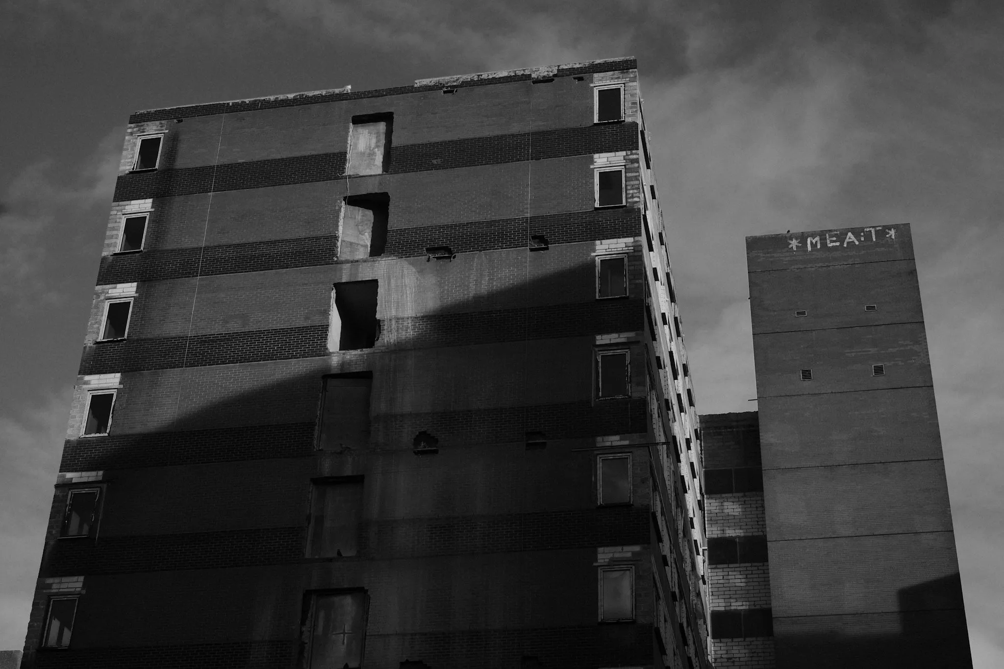 A derelict brick tower block against a bright sky, windows knocked out and floors part-demolished, the word MEAT painted high on the lift shaft beside it.