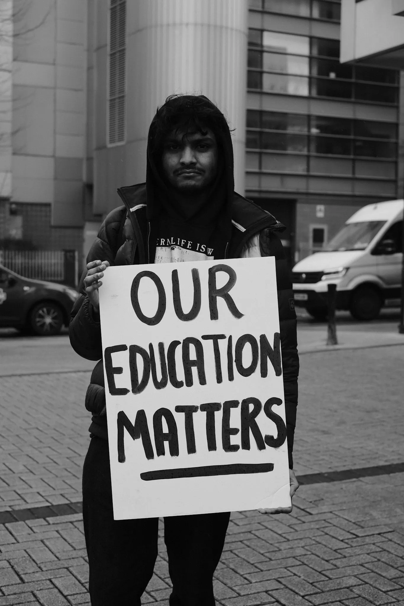 A student in a hooded coat stands on the pavement holding a hand-painted placard reading OUR EDUCATION MATTERS.