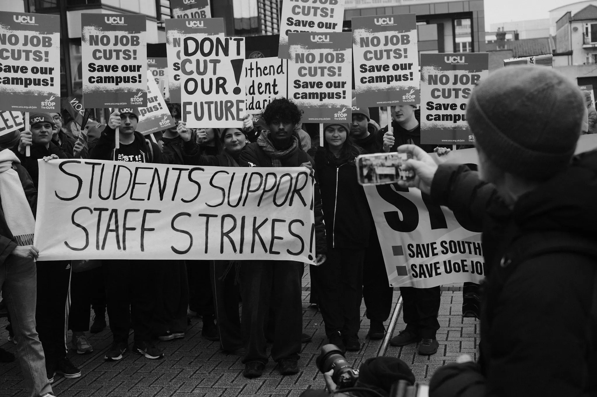 Students hold a hand-painted banner reading STUDENTS SUPPORT STAFF STRIKES beneath a forest of UCU No Job Cuts! Save our campus! placards, a phone raised to film them.