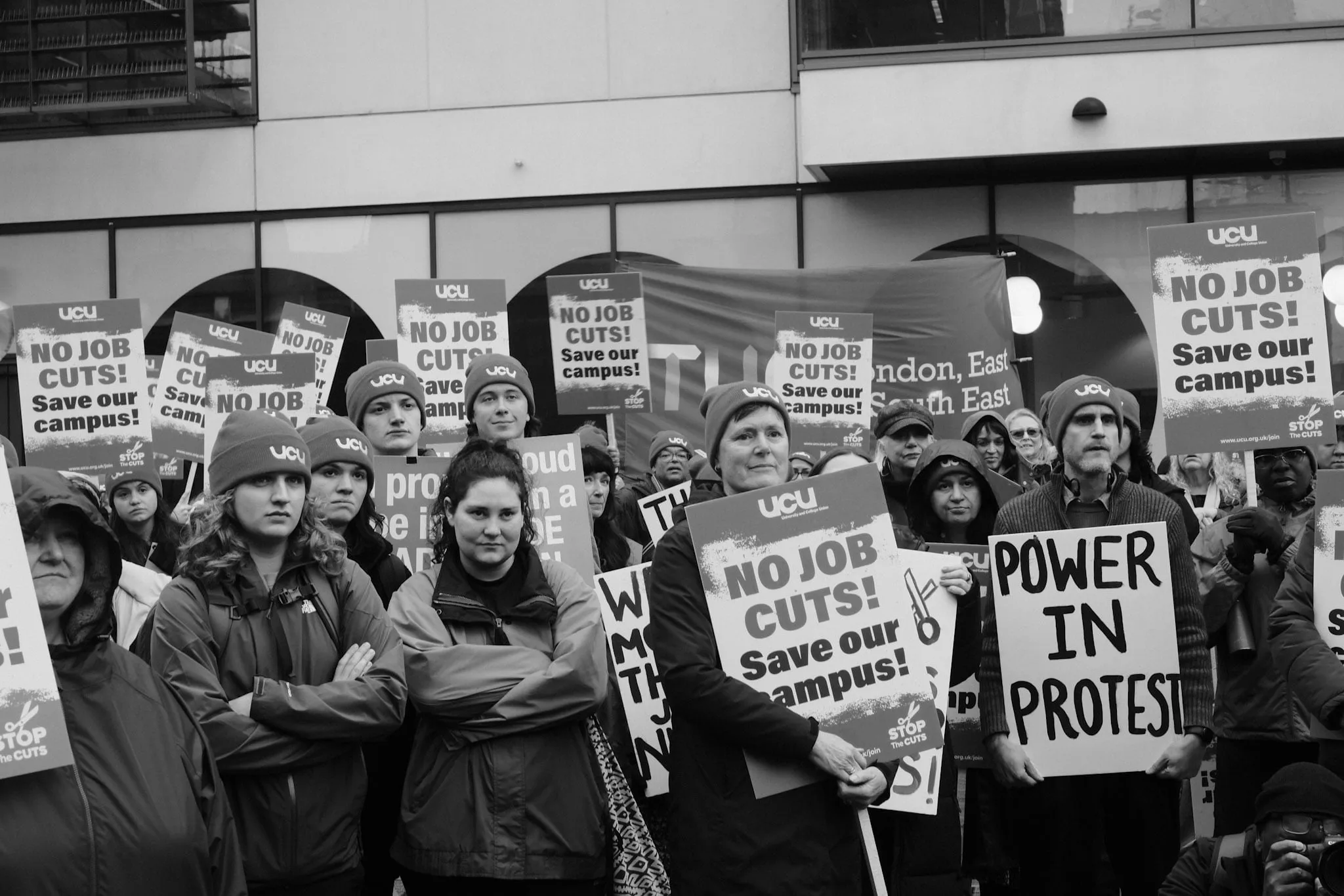 A dense crowd of pickets, most in UCU beanies, packed behind rows of No Job Cuts! placards and a homemade POWER IN PROTEST sign, a TUC regional banner strung up behind.