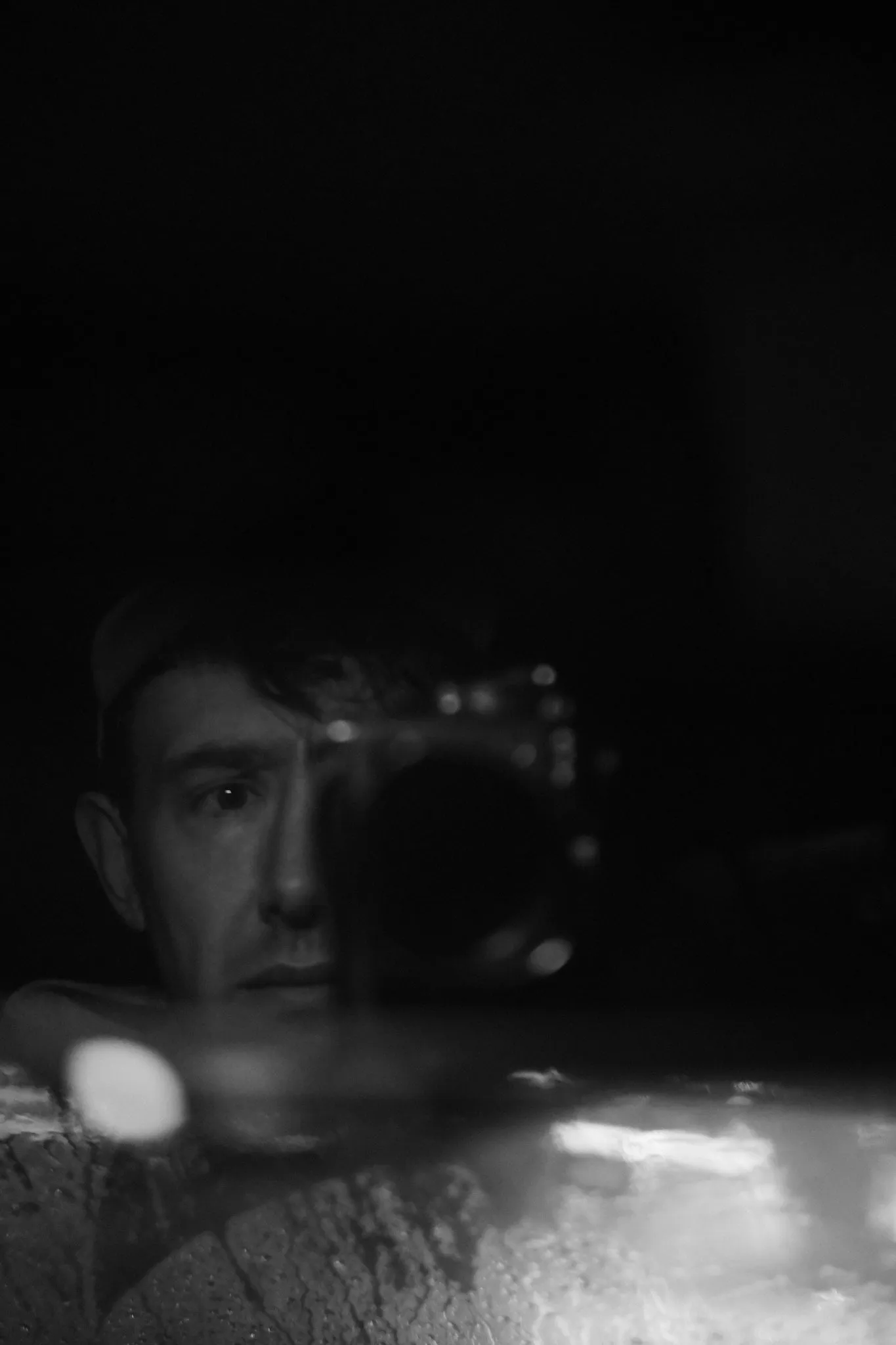 A photographer's reflection in dark glass, camera raised to one eye, most of the frame in shadow.