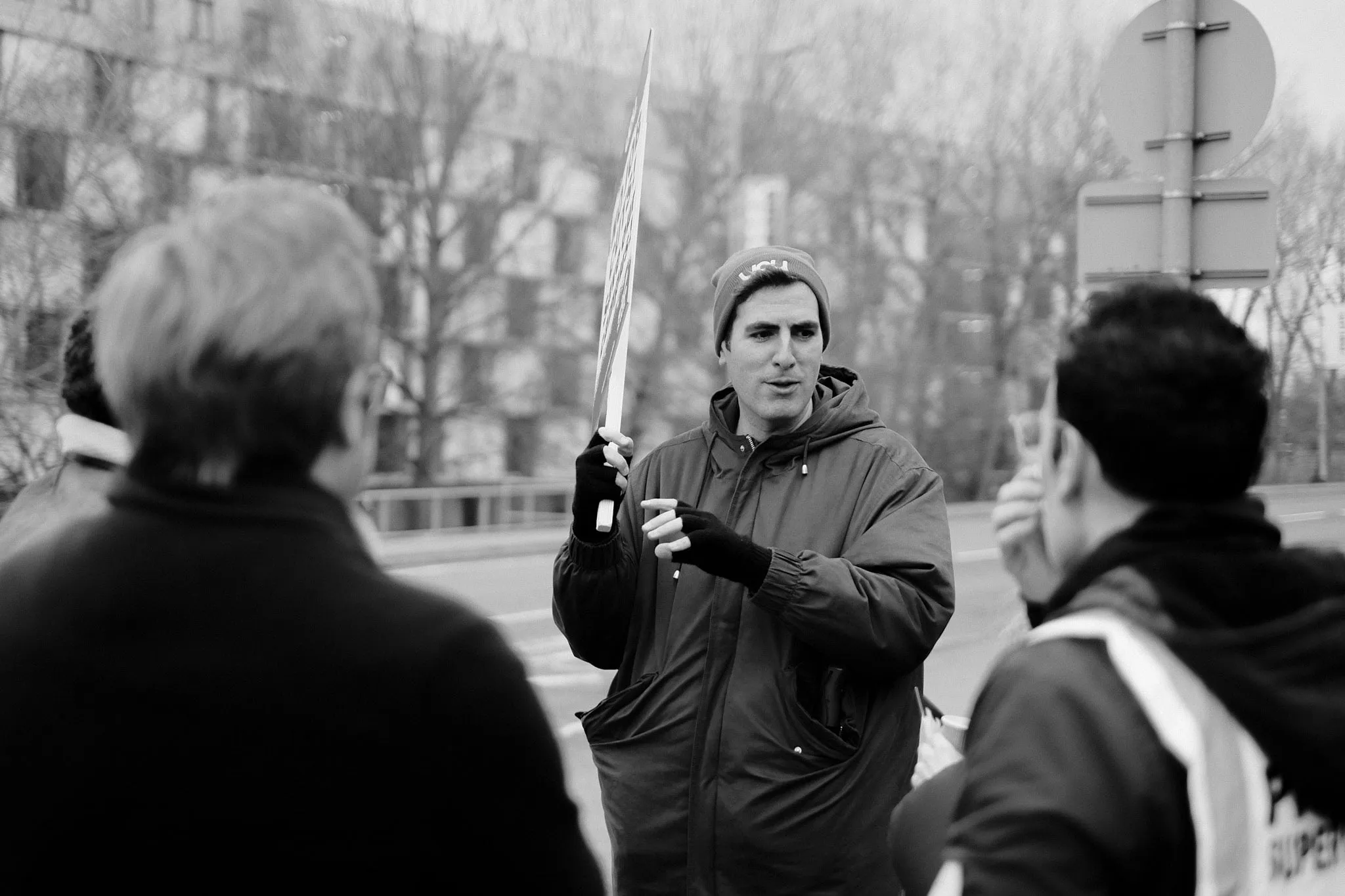 A picket in a UCU beanie holds a placard aloft and talks to two others on a roadside pavement, campus buildings and bare trees behind.