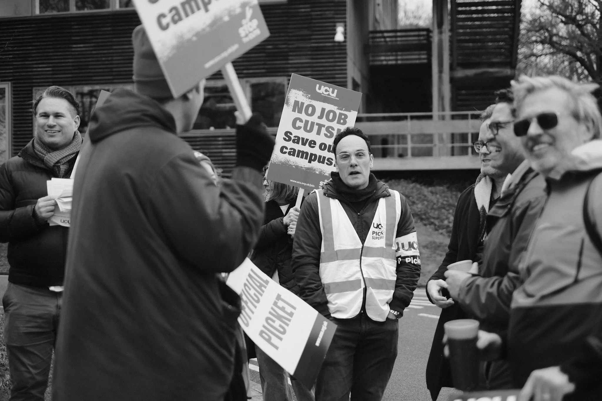 A UCU picket supervisor in a hi-vis vest stands mid-conversation on the picket line, an OFFICIAL PICKET placard and a No Job Cuts! board raised around him.