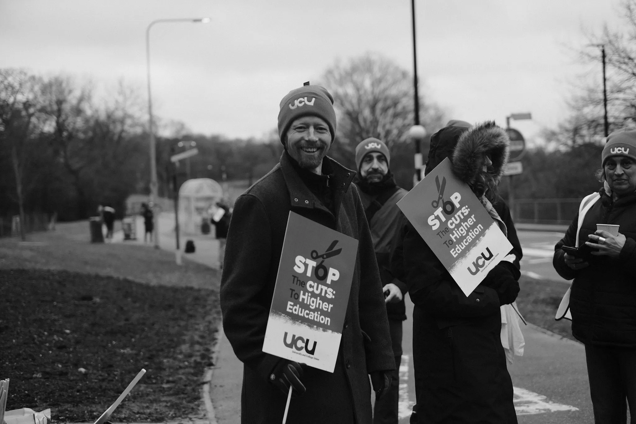 A picket in a UCU beanie grins at the camera holding a Stop the Cuts: To Higher Education placard on a roadside verge, more pickets behind him.