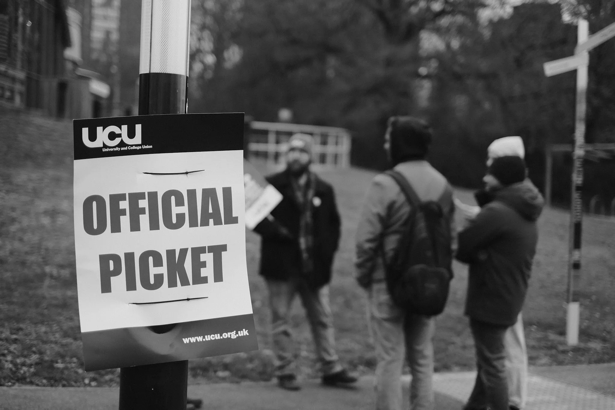 A UCU OFFICIAL PICKET notice cable-tied to a lamppost, three figures blurred on the path beyond.
