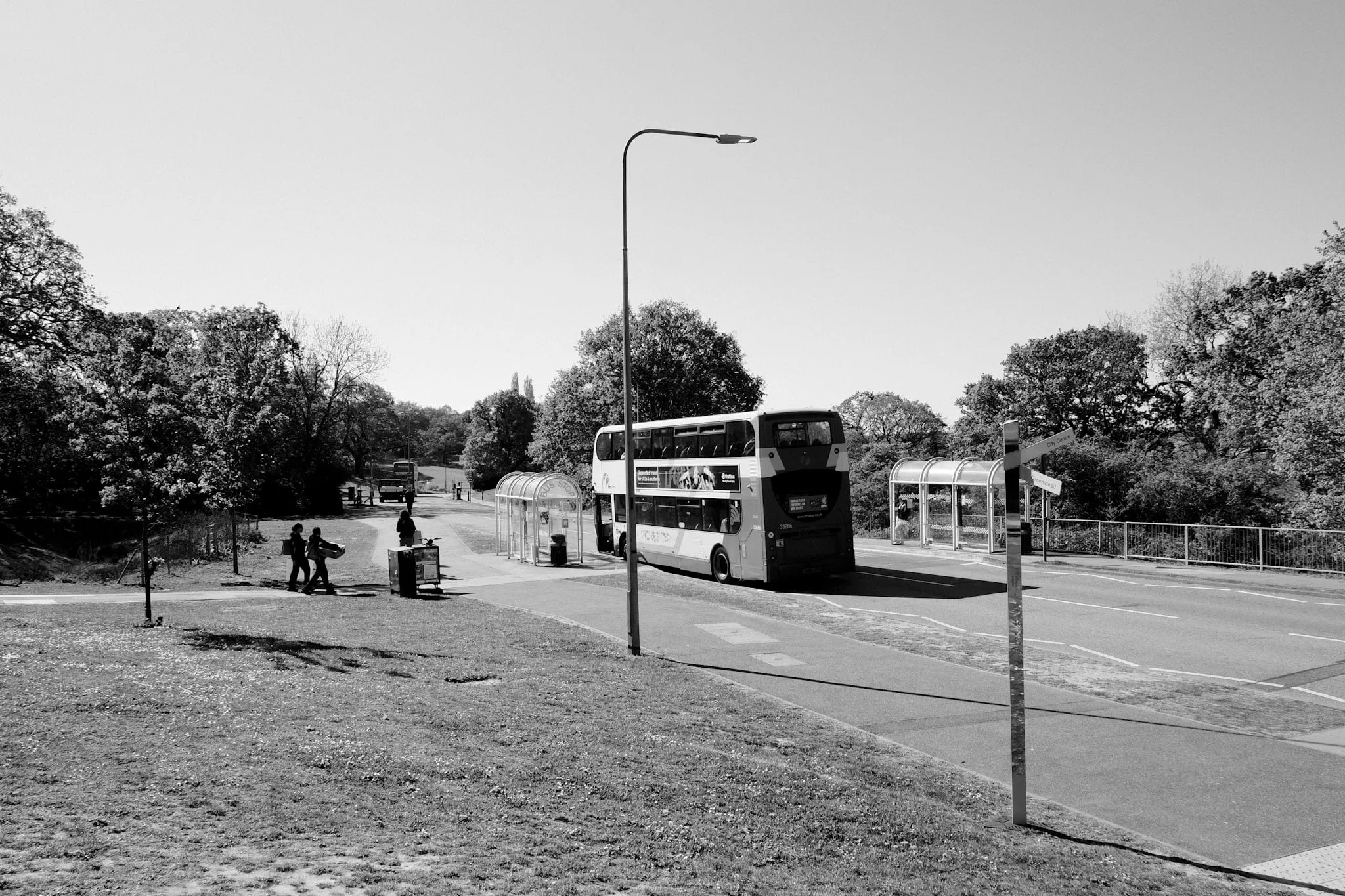 An empty campus road in bright summer sun, a single double-decker pulled in at a bus stop and a few people walking the path beside it.