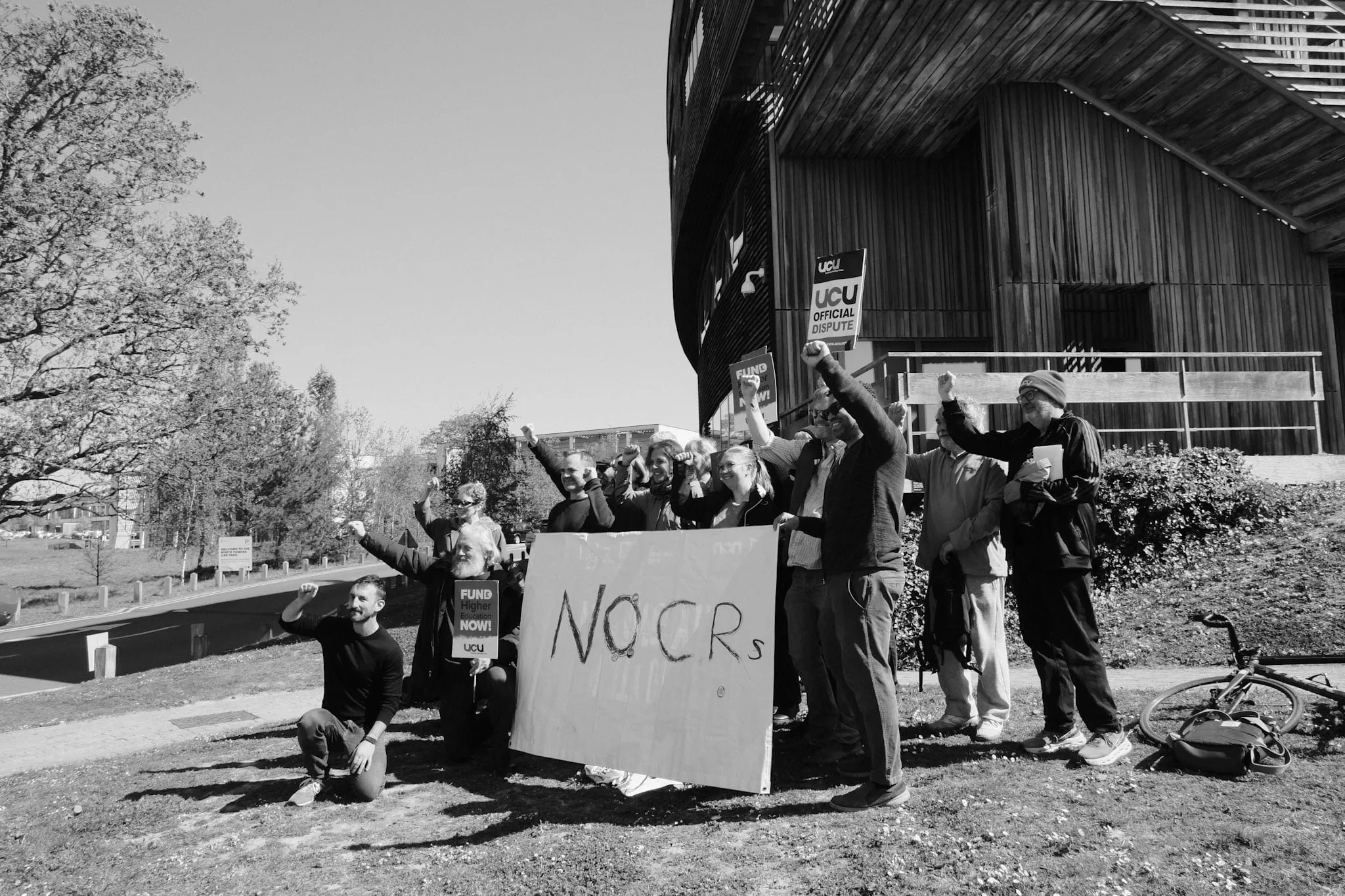A dozen people outside a timber-clad campus building raise their fists behind a hand-painted NO CRs banner in bright sun, a UCU Official Dispute placard beside them.