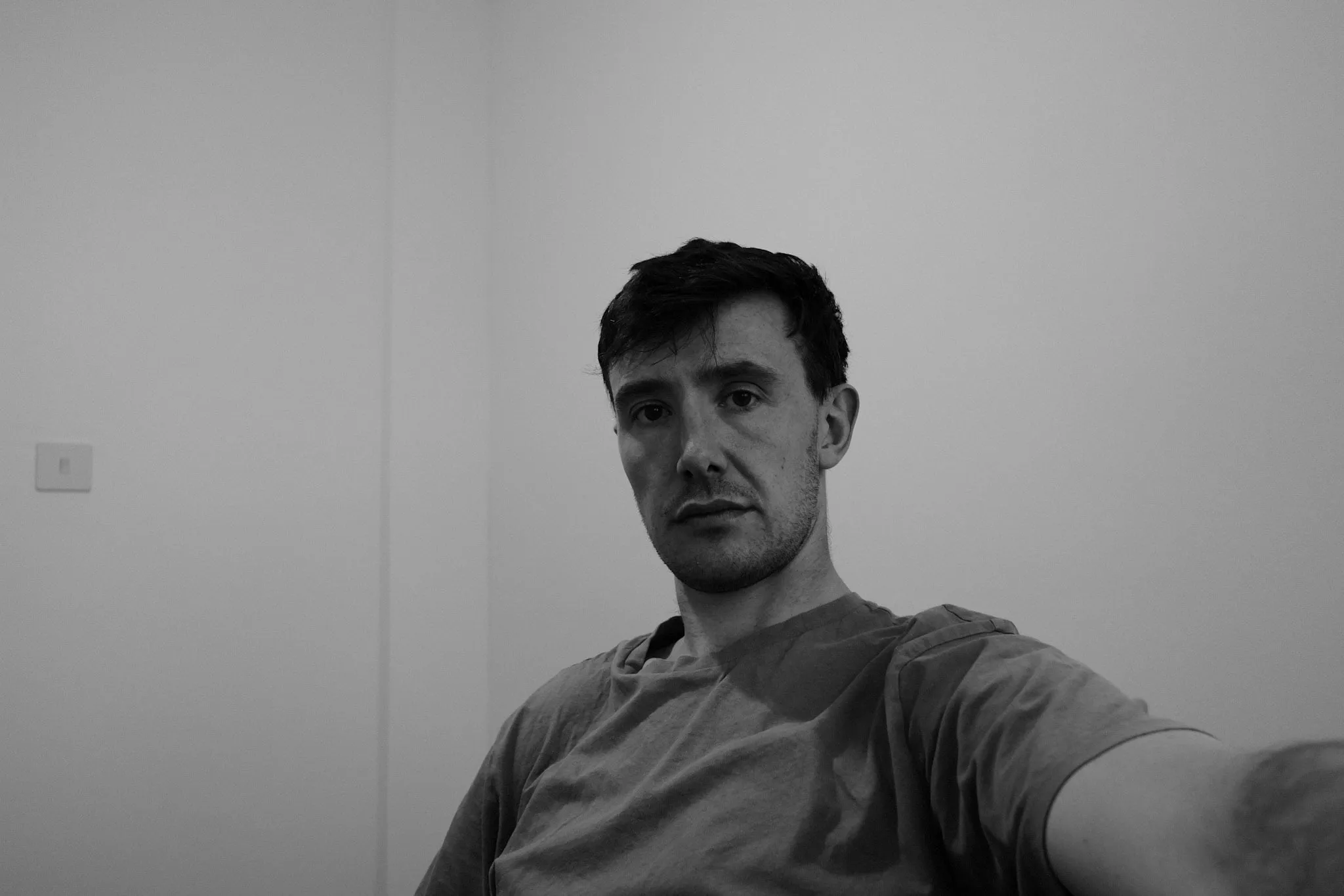 A man sits against a plain white wall in a t-shirt, one arm outstretched towards the camera, looking straight into it.