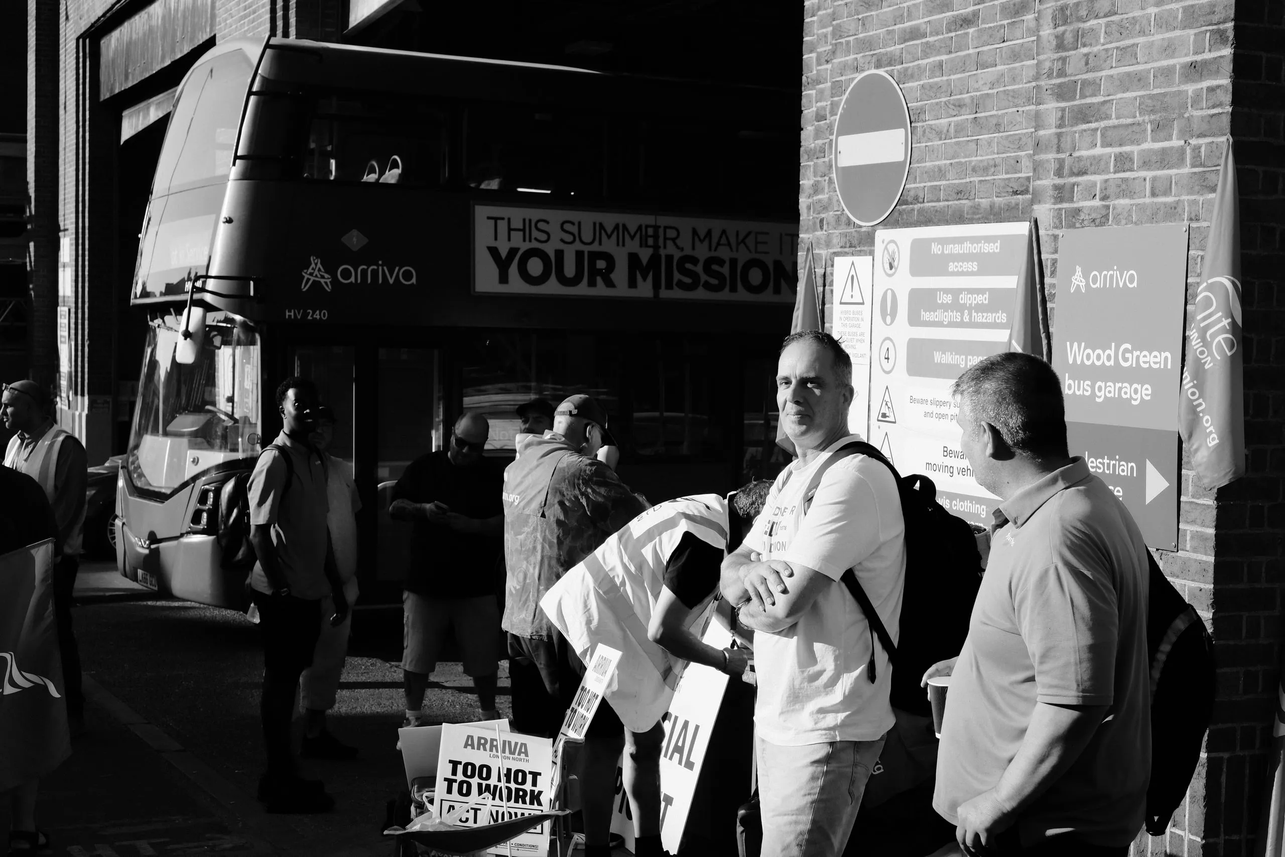 Pickets outside Wood Green bus garage in early morning sun, an Arriva double-decker behind them and Too Hot to Work placards at their feet.