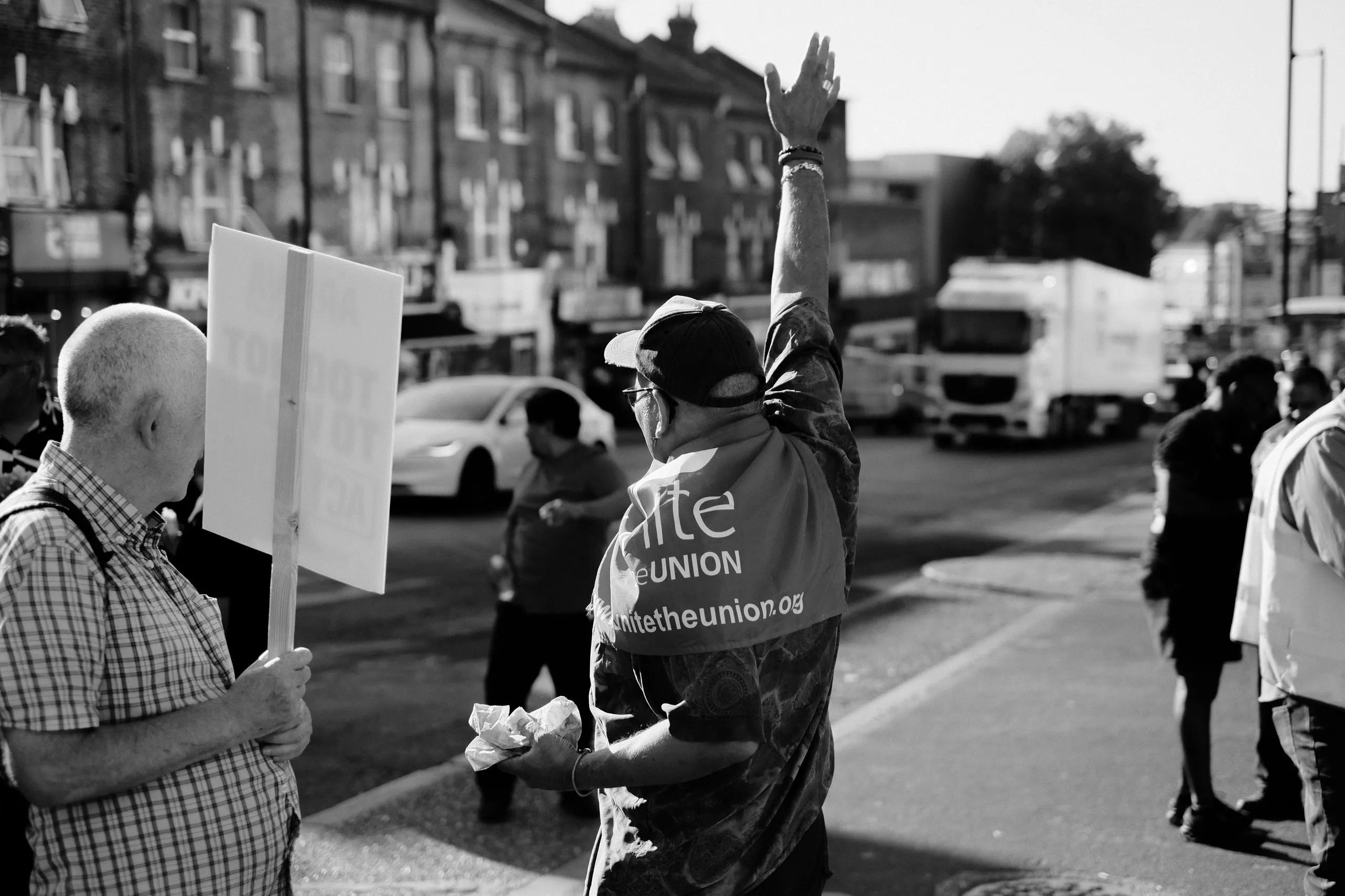 A picket with a Unite flag over his shoulder raises an arm to passing traffic on the main road, another picket beside him holding a placard.