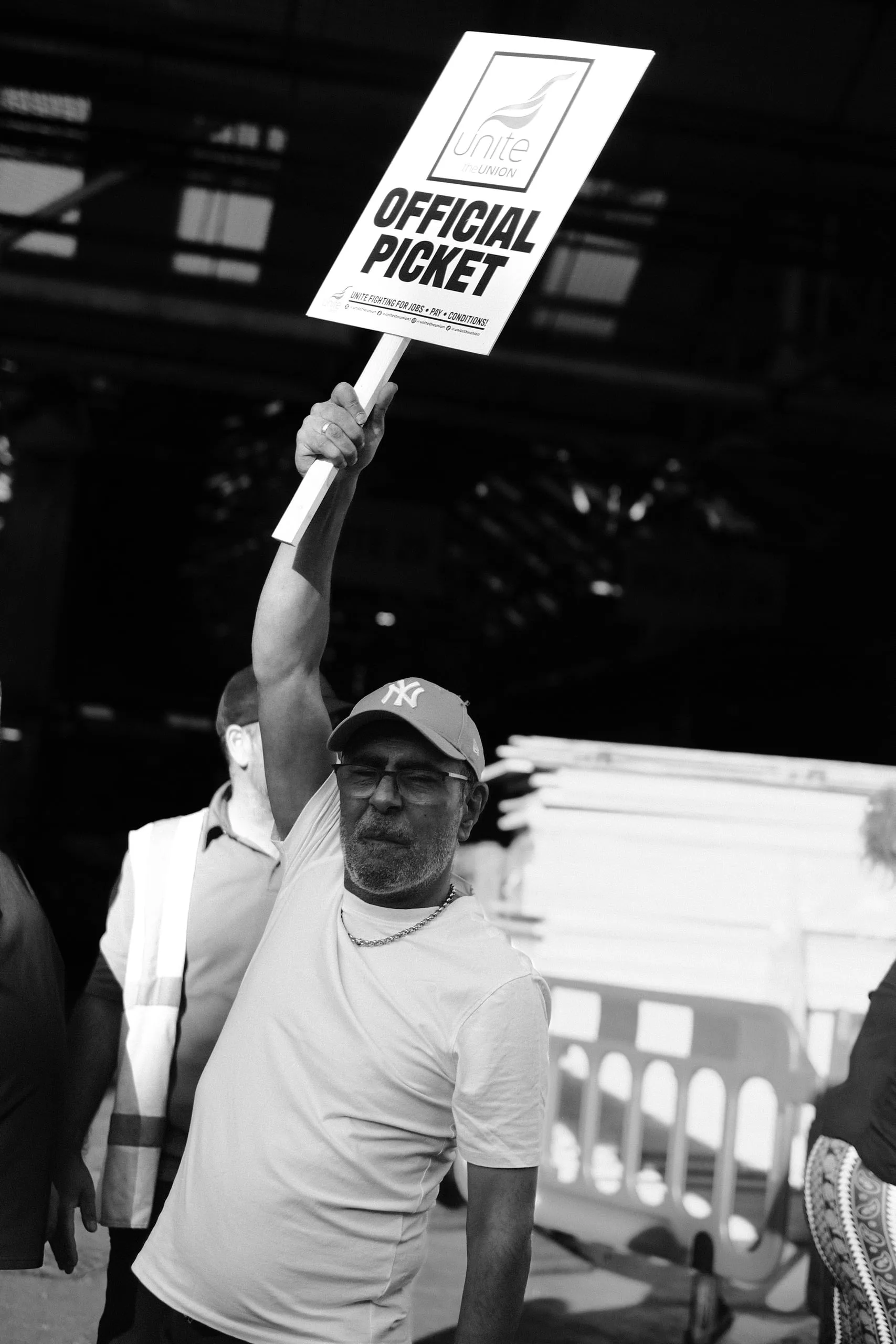 A picket in a white t-shirt and cap holds a Unite Official Picket placard above his head.