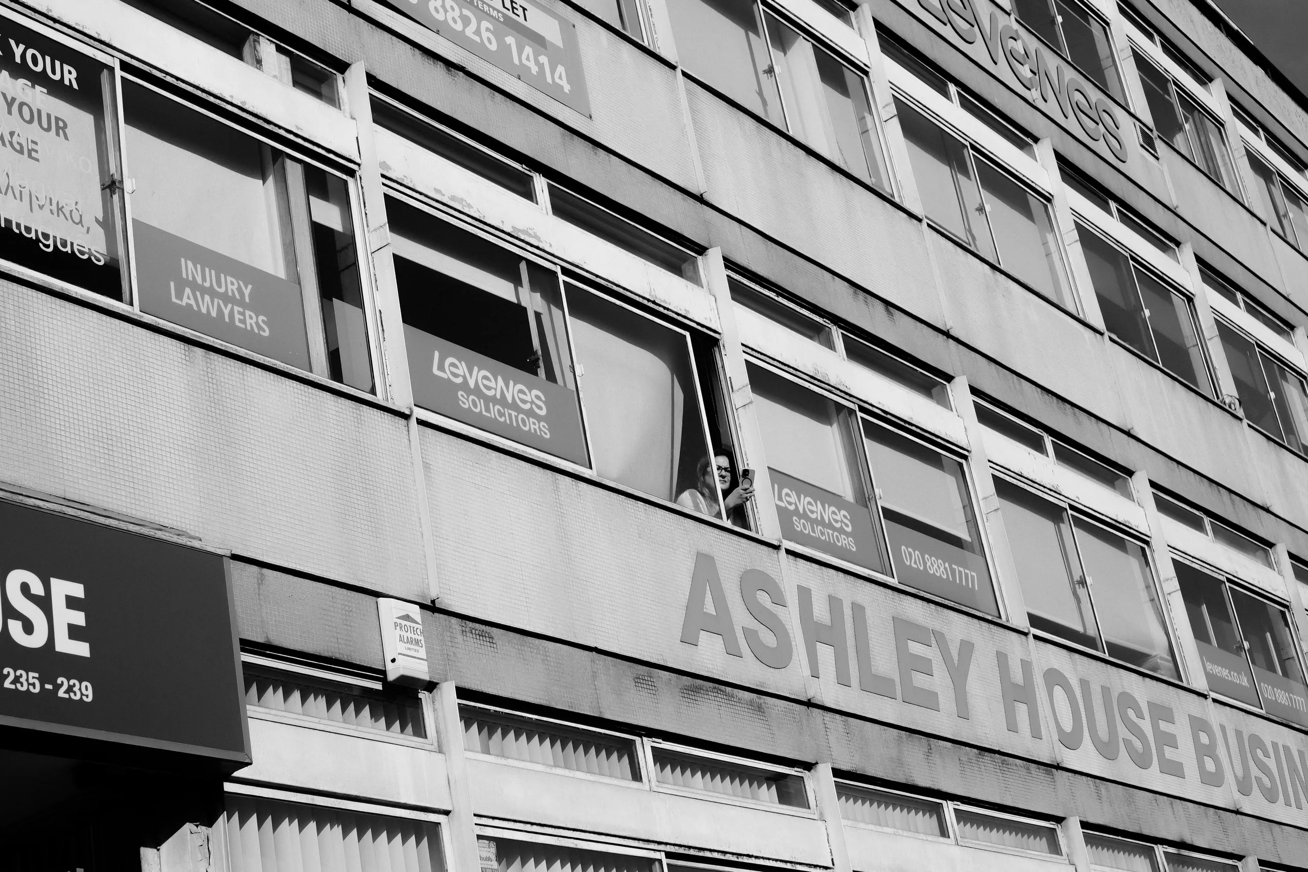 A woman leans out of an upper window of an office block opposite, filming the picket on her phone.