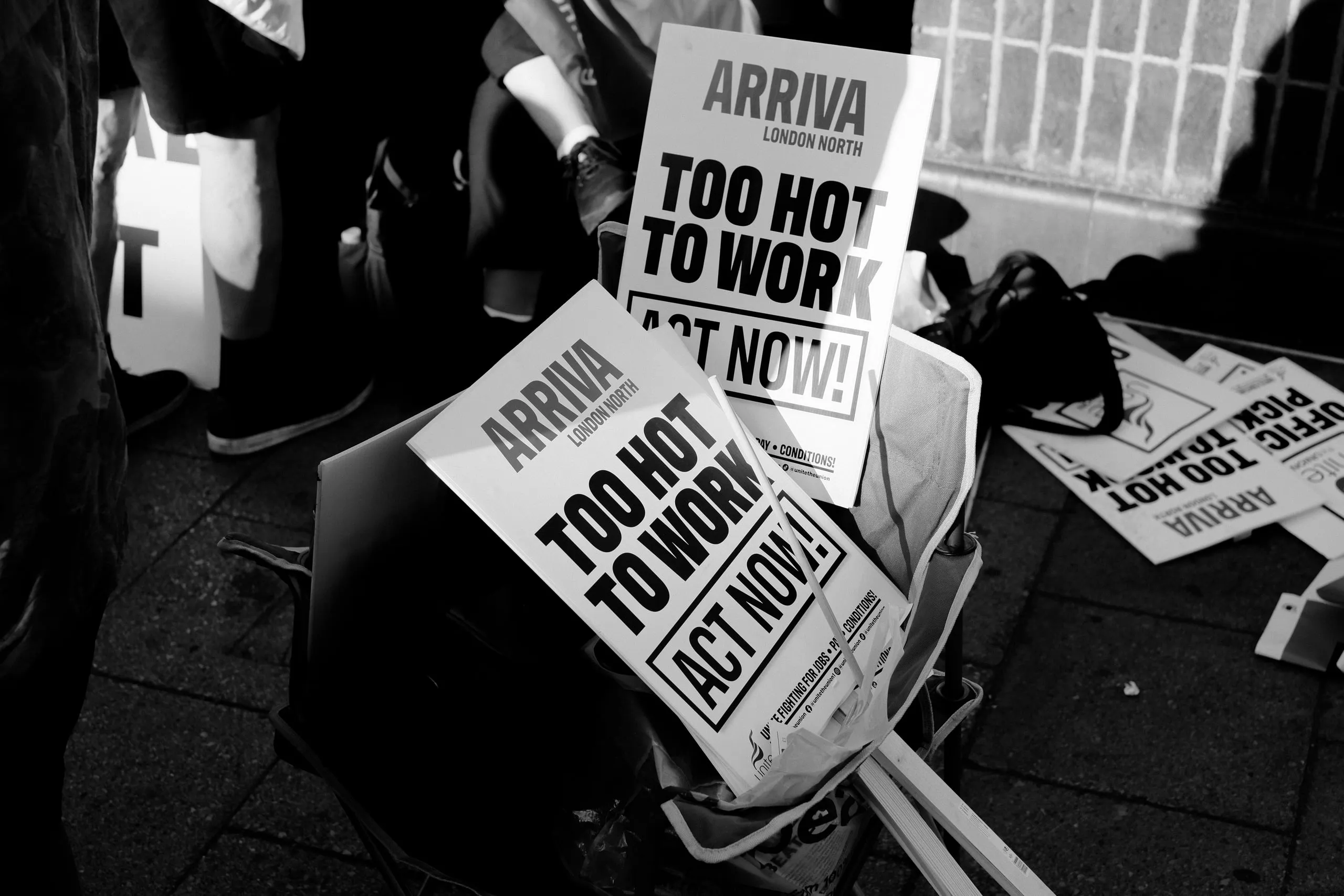 A stack of Arriva London North “Too Hot to Work — Act Now!” placards in a bag on the pavement, more scattered beside it.