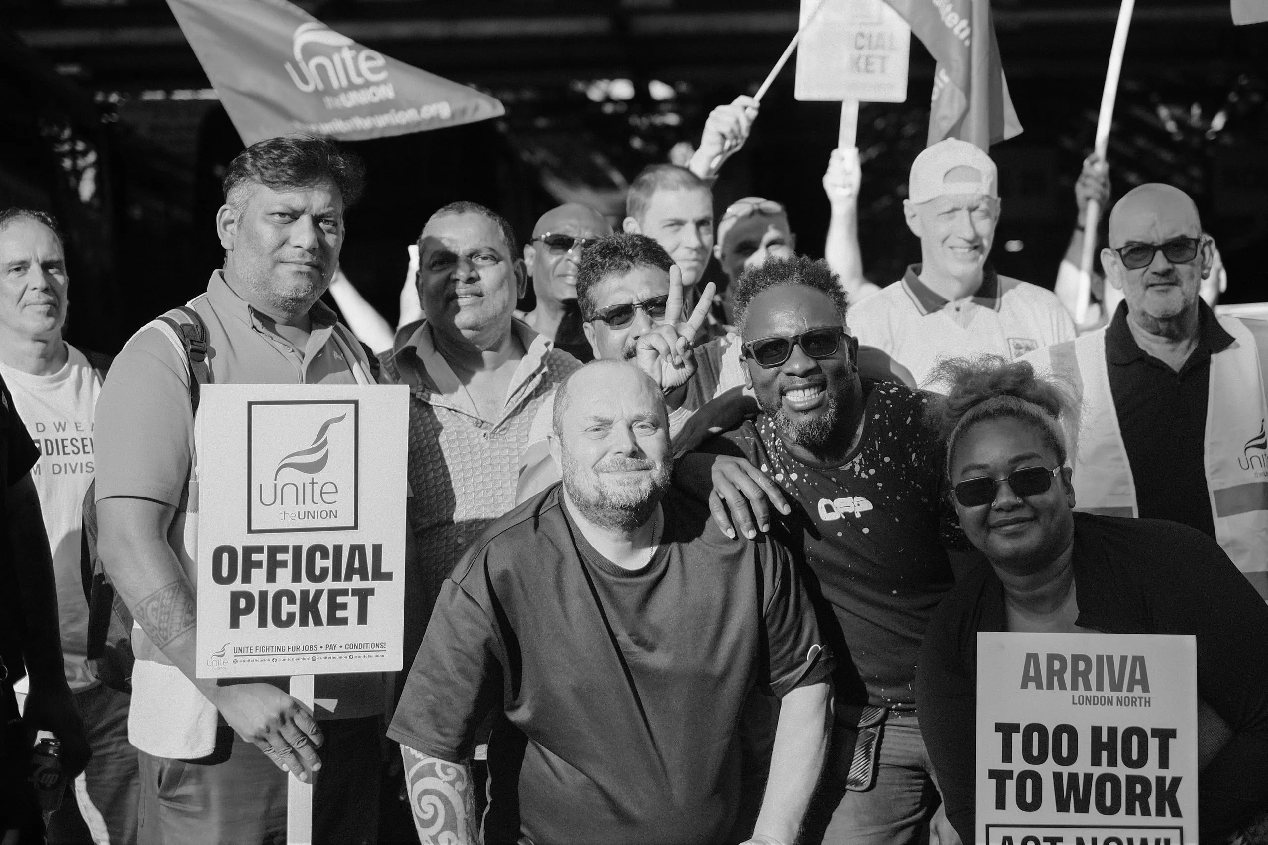 A large group of pickets crowded together for a photograph, Unite flags and Official Picket placards raised, several smiling at the camera.