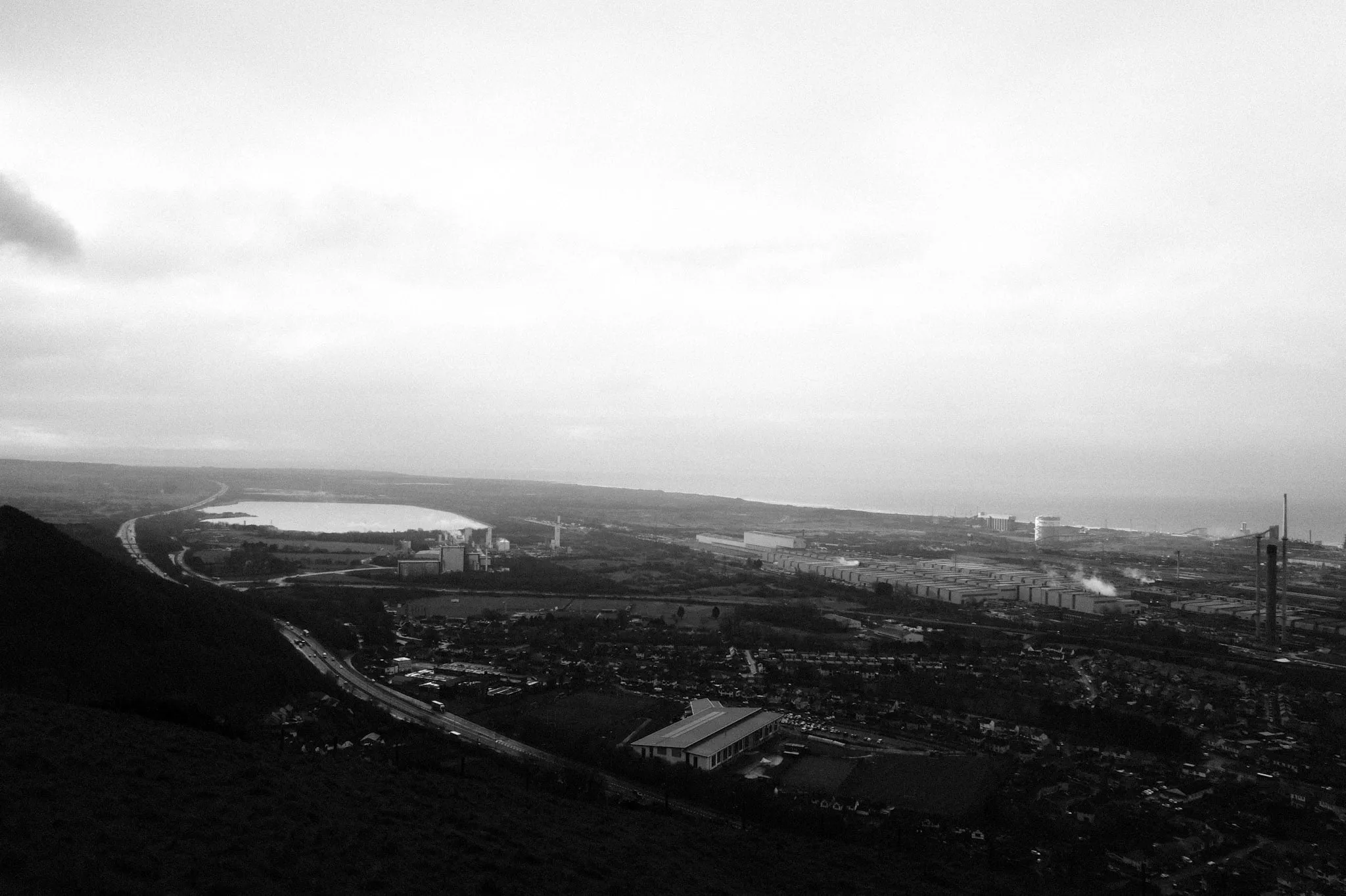 The valley from high on the hillside at dusk, a motorway curving past a reservoir towards the coast, industrial sheds and works stretching along the shoreline into haze.
