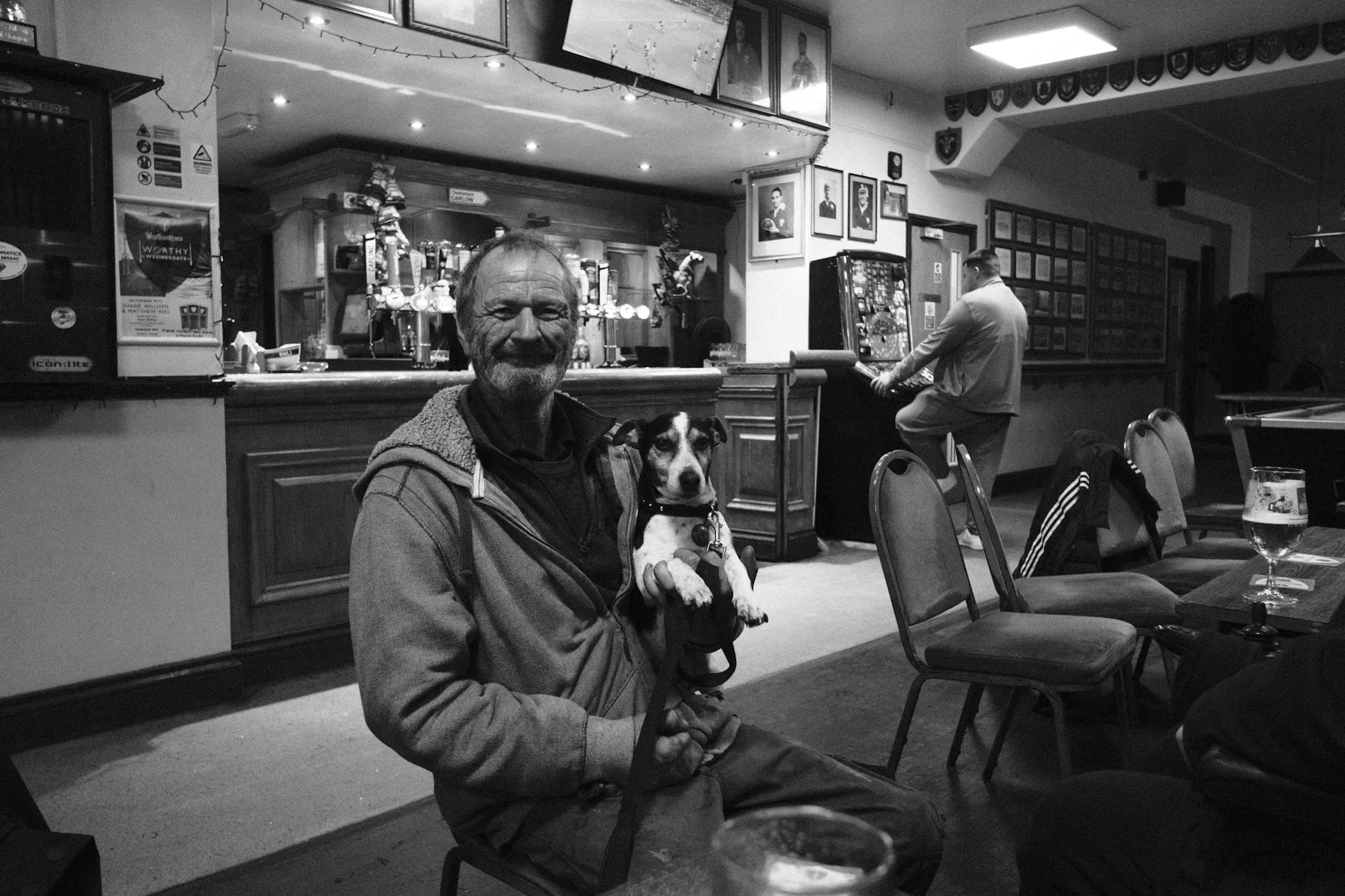A bearded man in a fleece-lined hoodie sits in a club holding a Jack Russell terrier on his lap, the bar and its beer taps behind him and another man at a fruit machine.