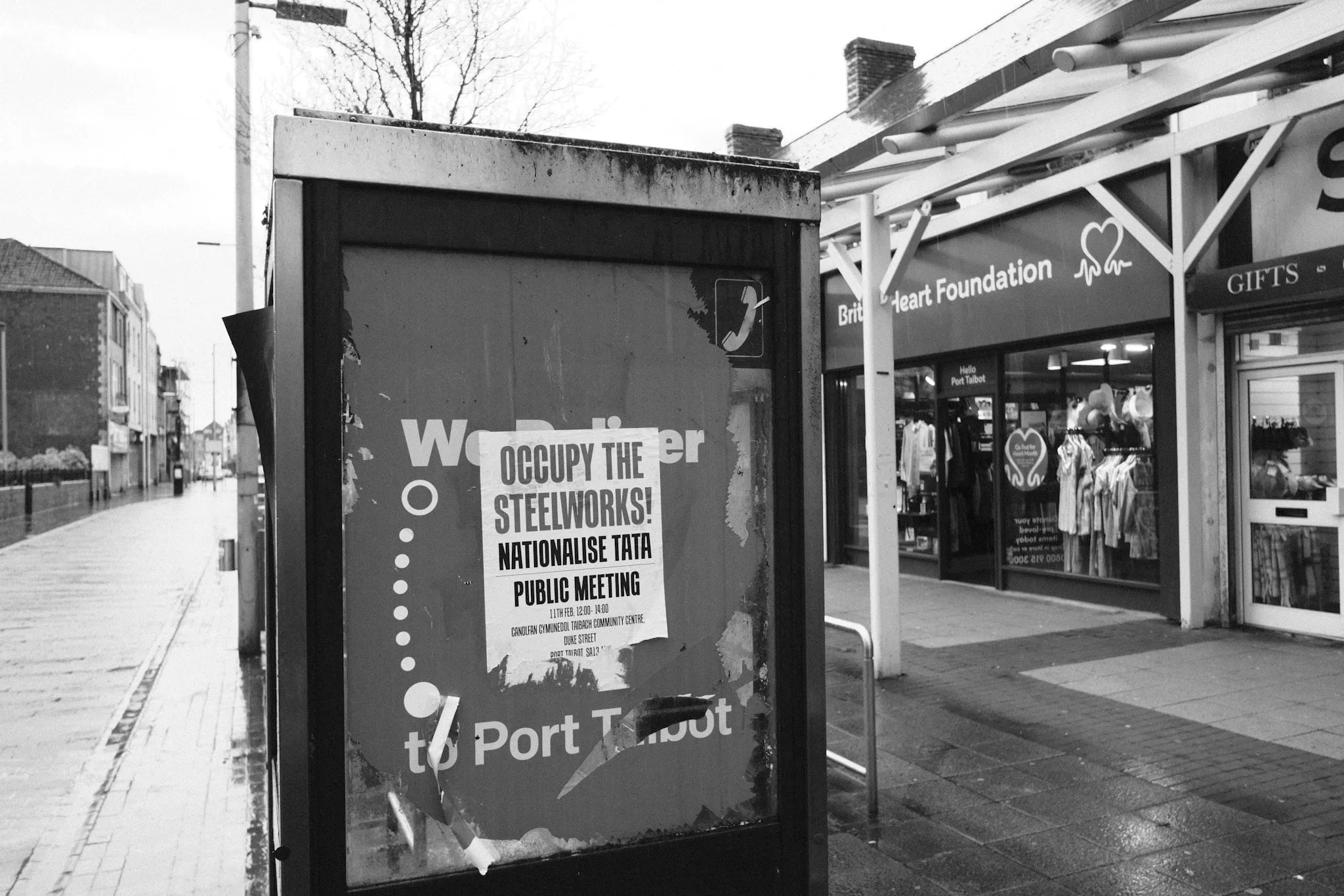 A flyposted sheet reading “Occupy the Steelworks! Nationalise Tata”, advertising a public meeting, pasted over a torn advert in a bus shelter on a wet high street, a British Heart Foundation shop behind.
