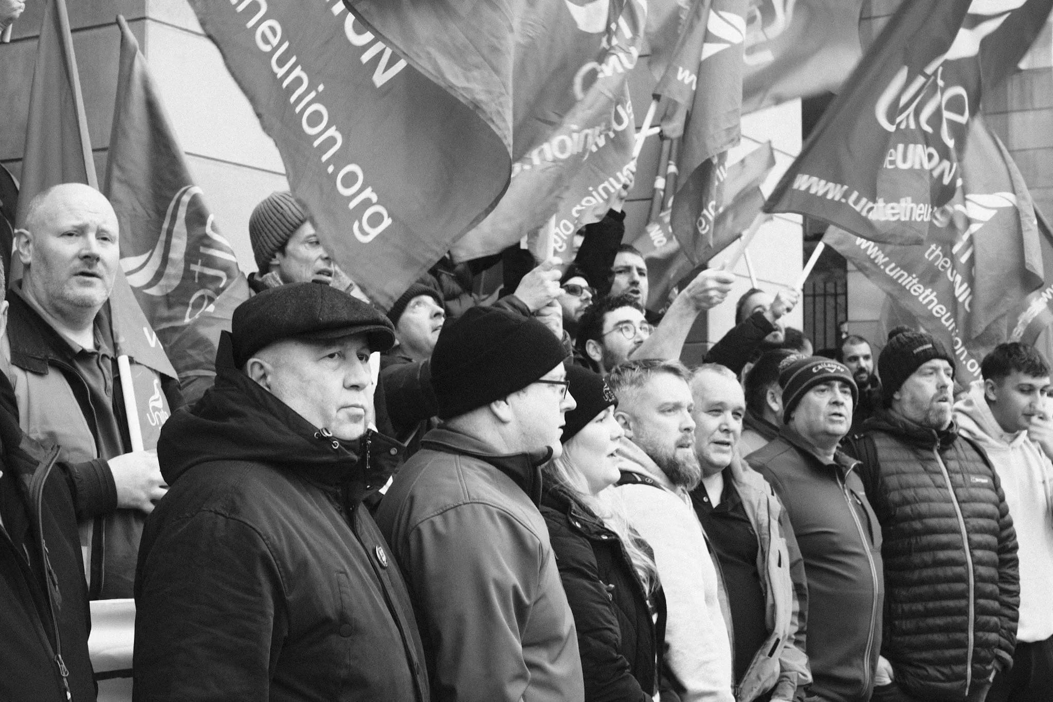 A packed crowd in winter coats and hats at a rally, Unite the Union flags raised and overlapping above their heads, several of them shouting.