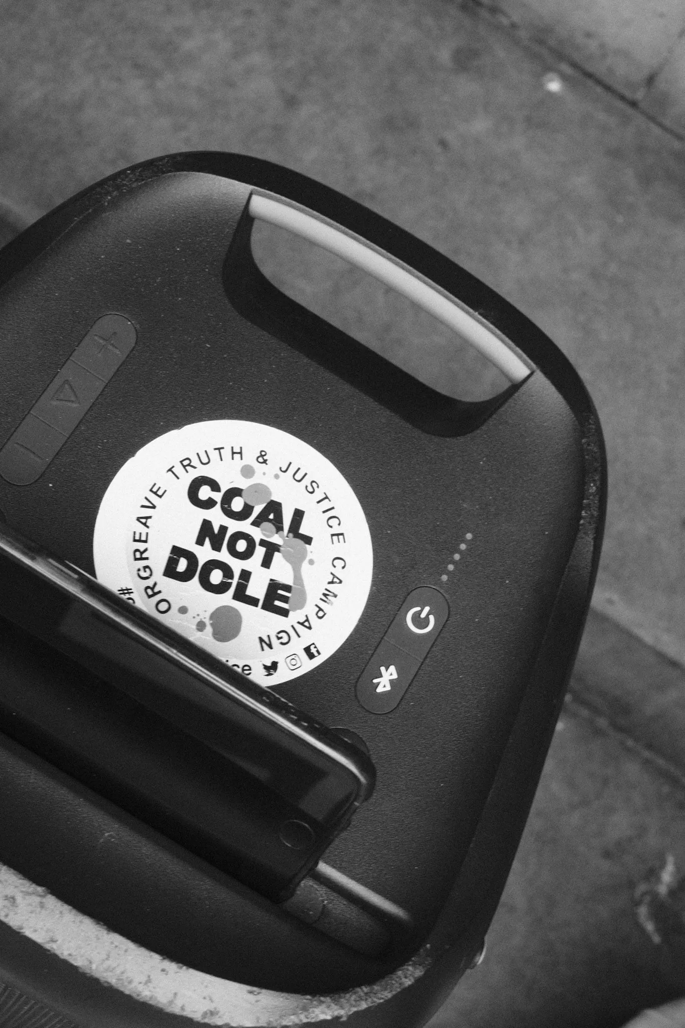 A round sticker reading “Coal Not Dole — Orgreave Truth & Justice Campaign” stuck to the top of a black portable speaker, a phone resting against it on the pavement.