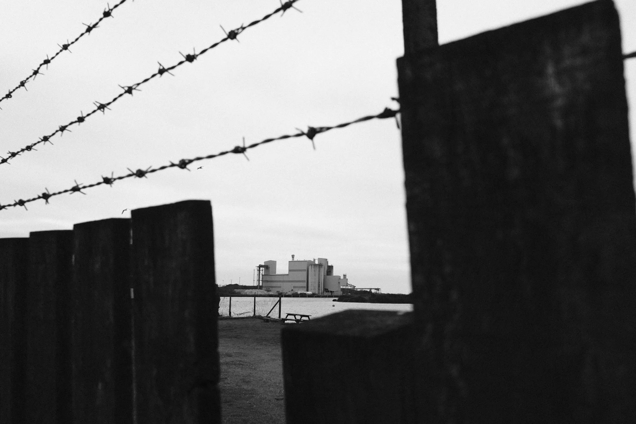 A pale industrial building across a stretch of water, seen between the silhouetted posts of a timber fence with barbed wire strung above.