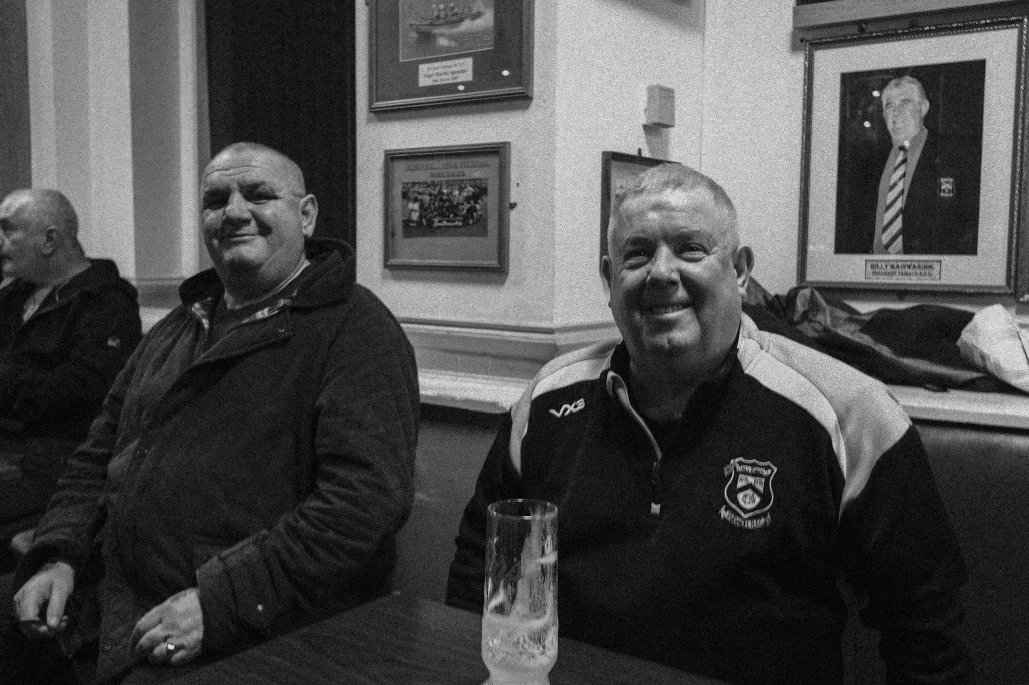 Two men sit smiling at a club table beside a half-drunk pint, framed rugby photographs and a club president’s portrait on the wall behind them.