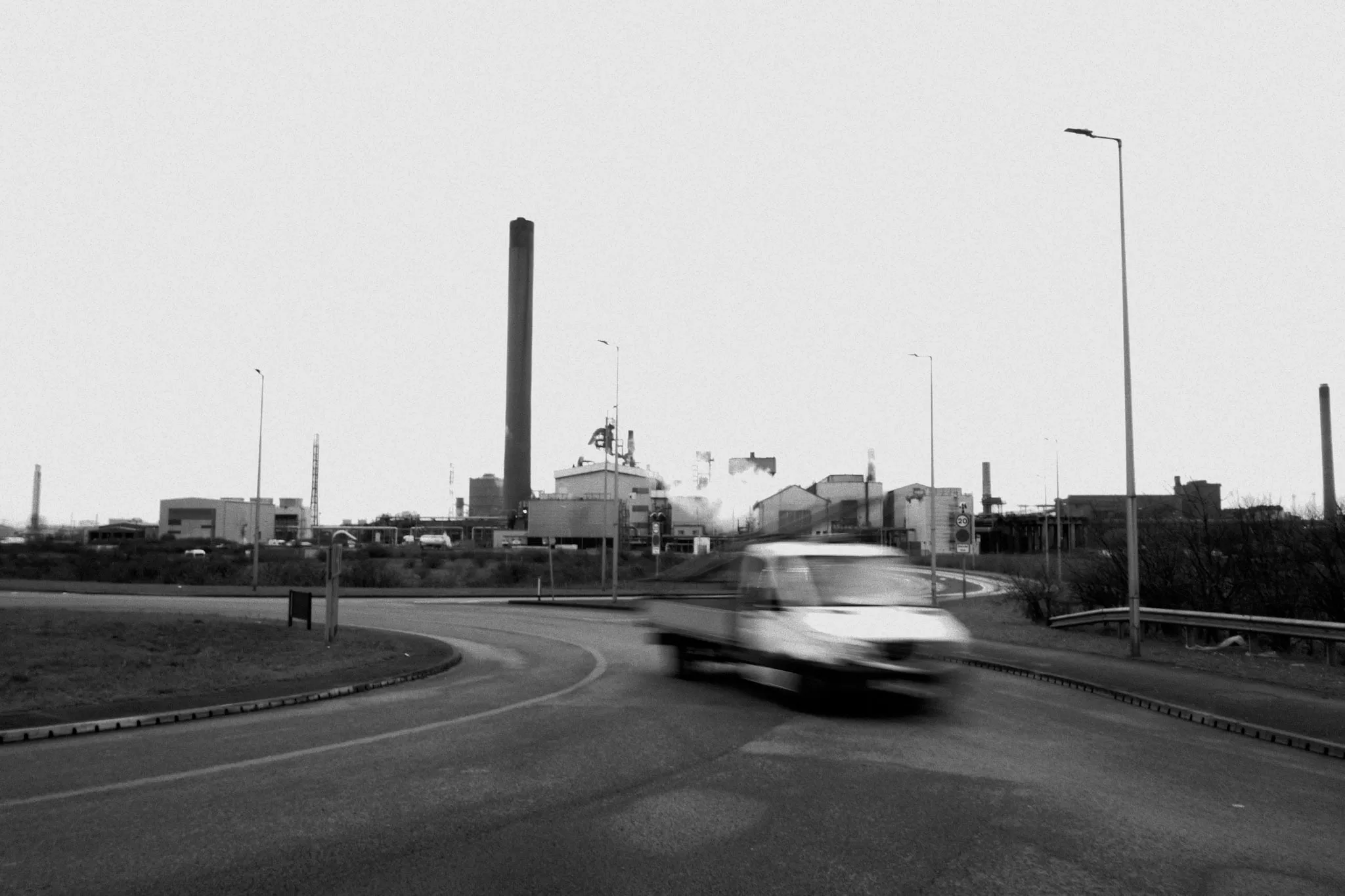 A van blurred with motion crossing a roundabout at dusk, the chimneys and sheds of the works standing flat against a pale sky behind it.