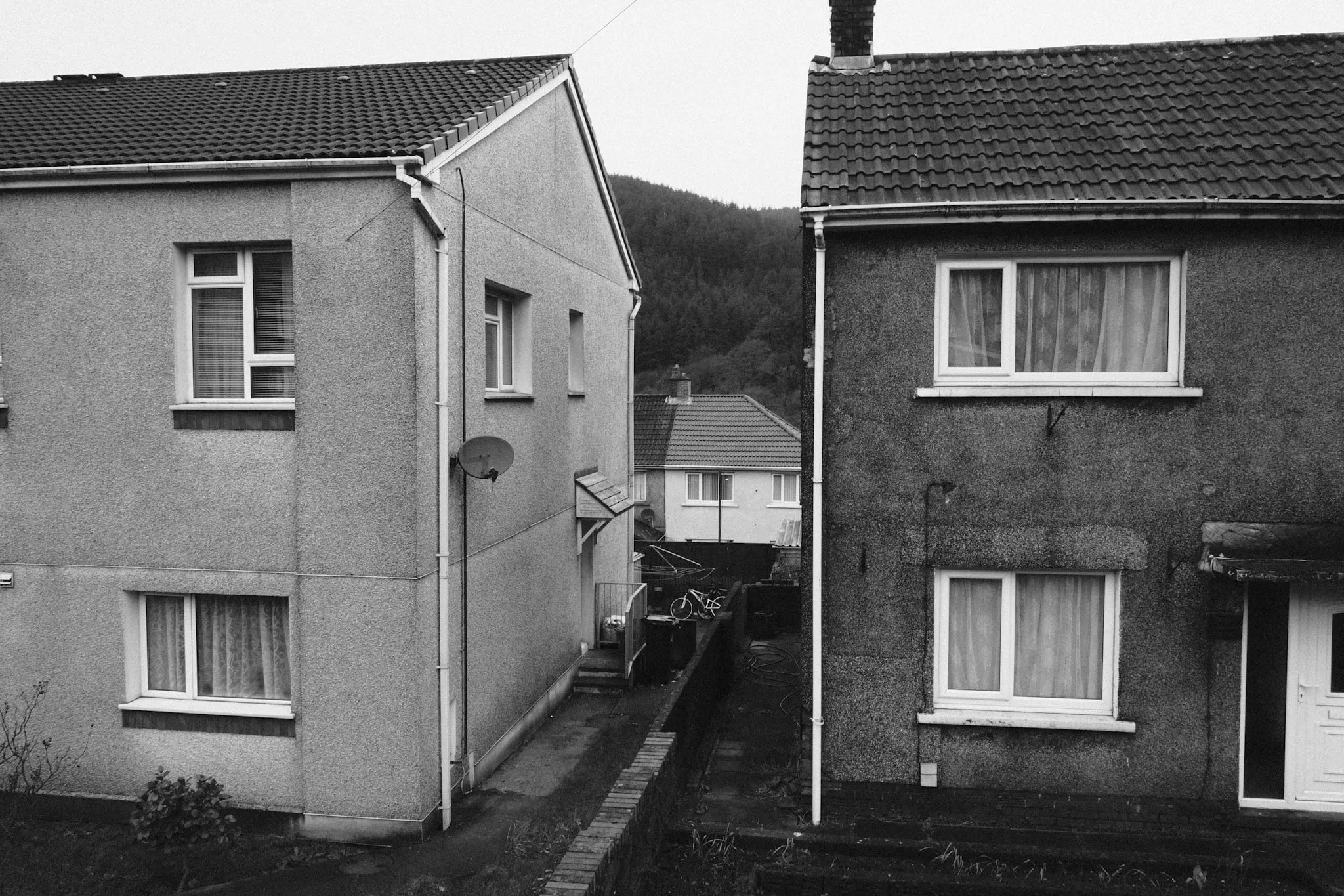 Two pebbledash houses seen end-on across a narrow alley, a satellite dish on one gable, a bicycle in the yard between them and a wooded hillside rising behind the roofs.