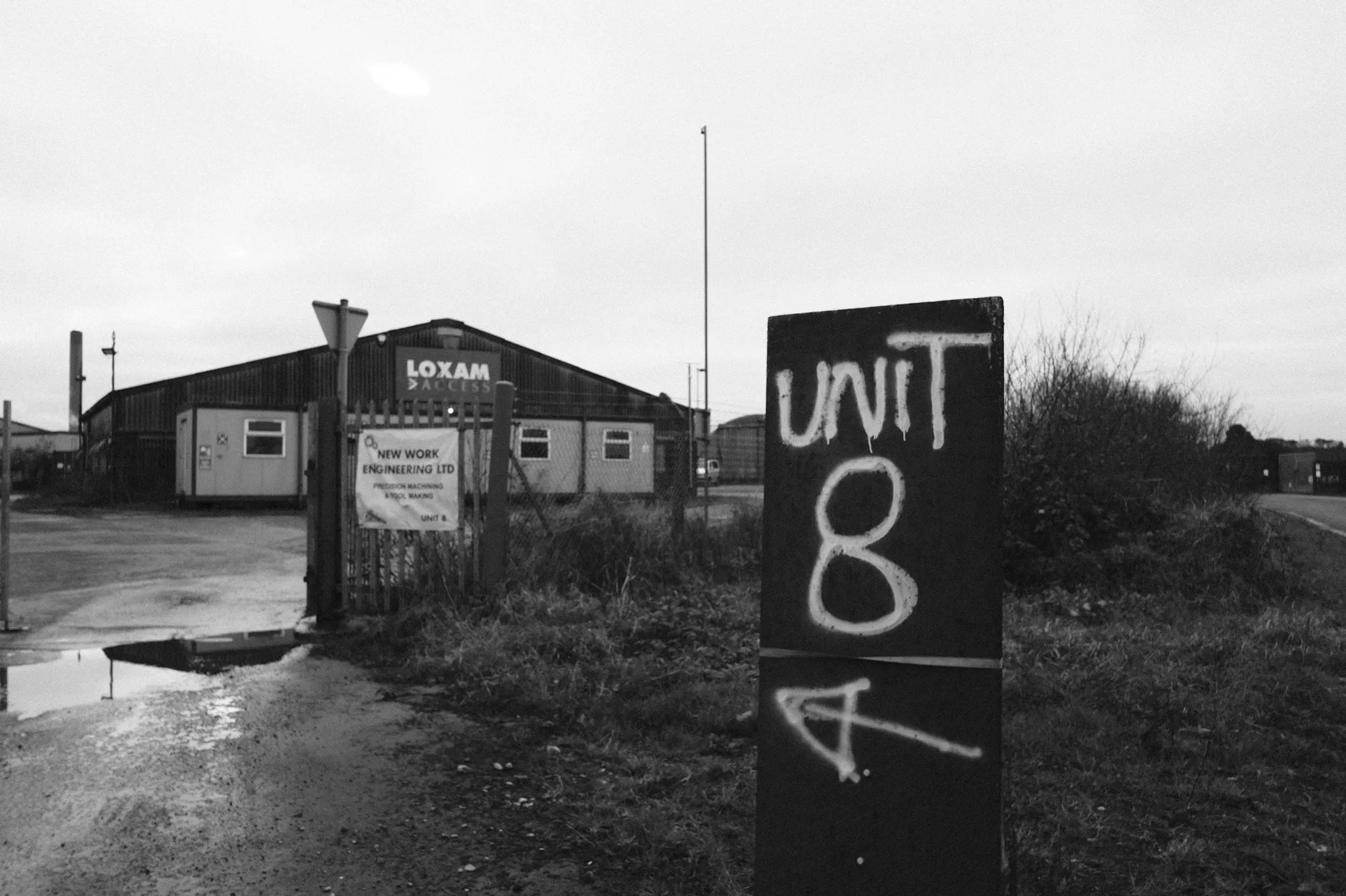 A hand-painted “Unit 8” sign on a black board at the entrance to an industrial estate, a corrugated shed and an engineering firm’s banner beyond the open gate, puddles across the track.