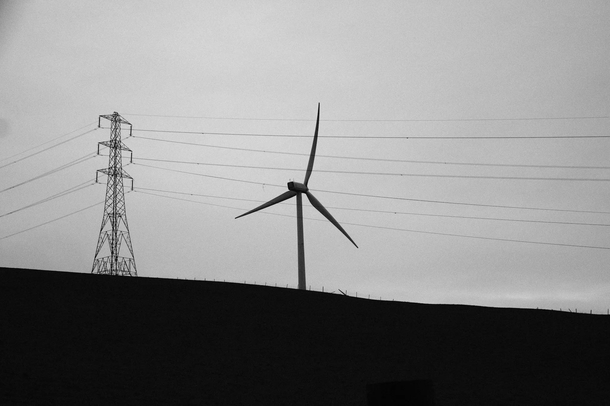 An electricity pylon and a single wind turbine on a dark ridge line against a flat grey sky, power lines running right across the frame.